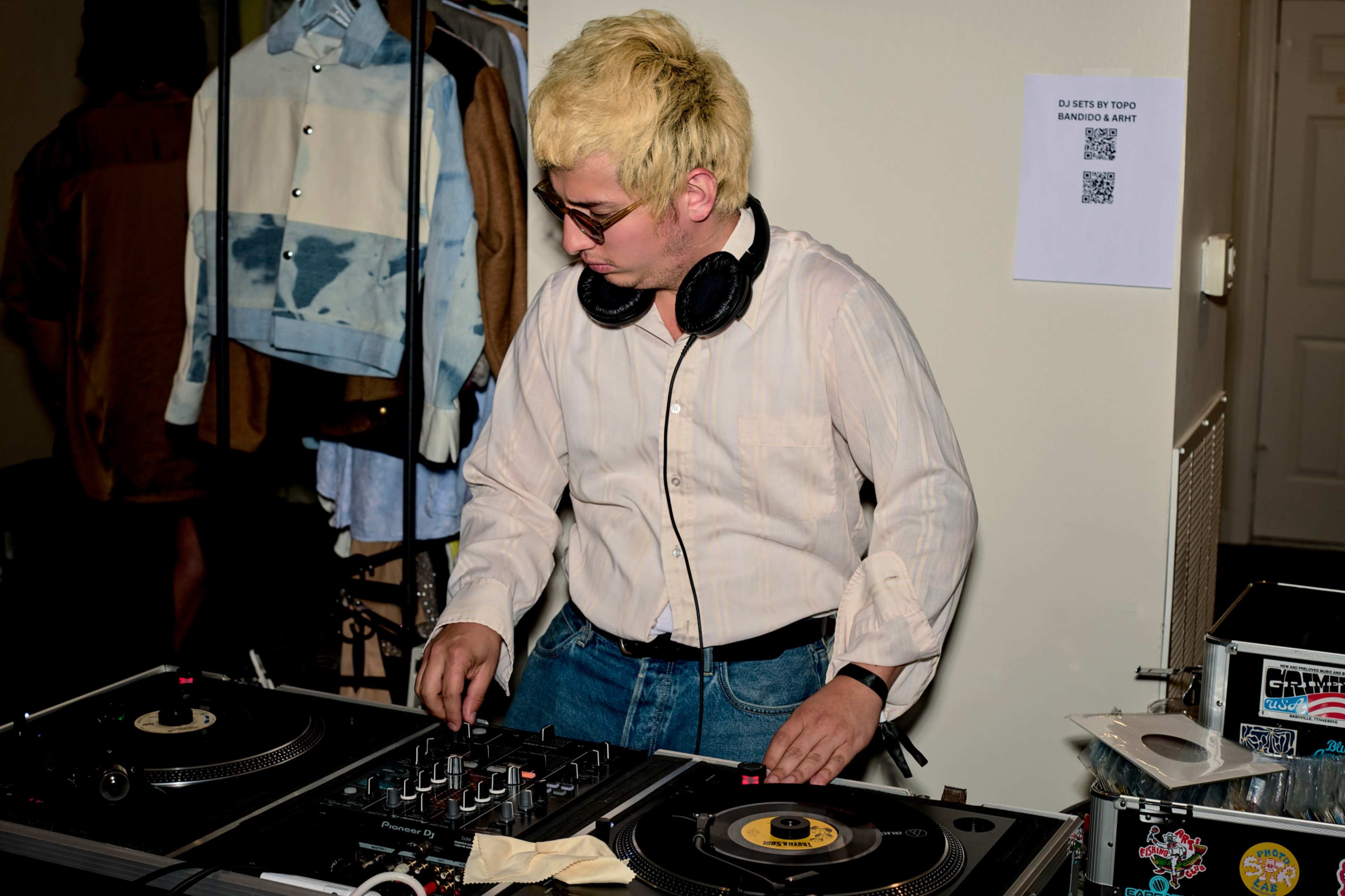 A DJ with blonde hair is mixing music at a turntable setup in a room with clothing hanging in the background.