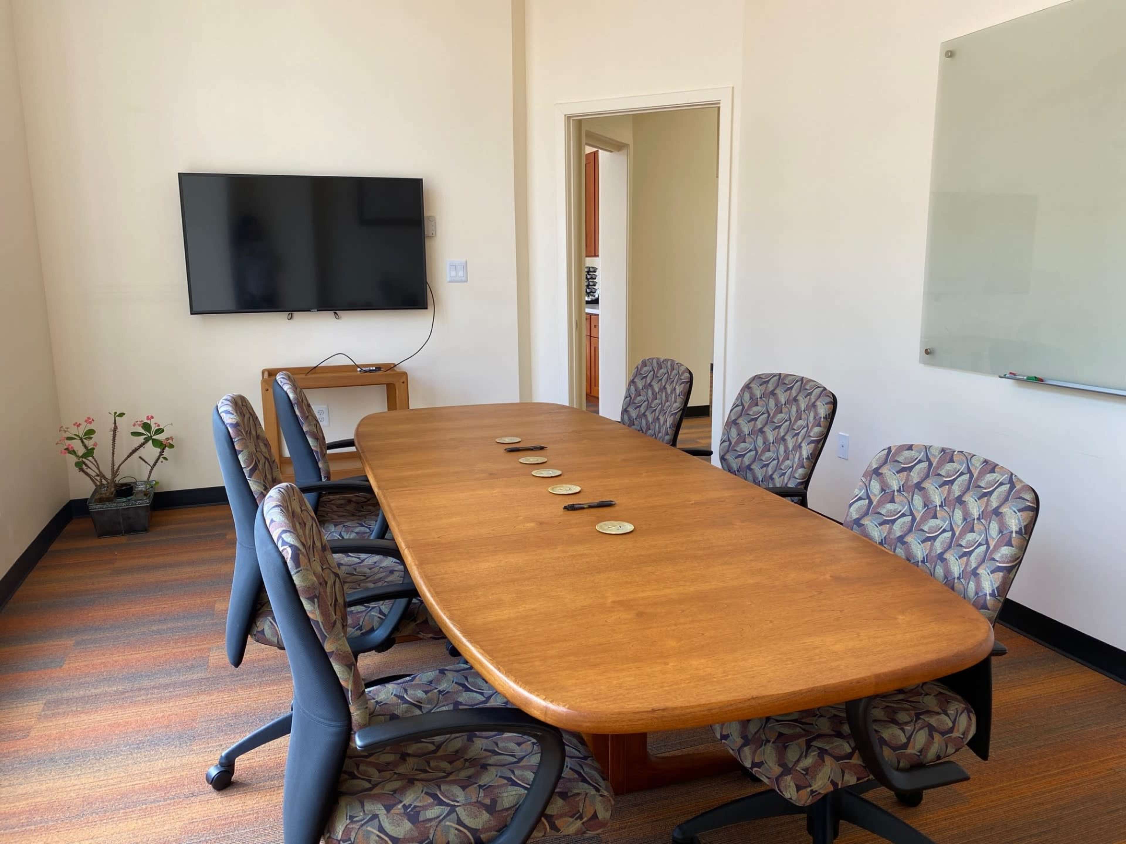 A conference room features a large wooden table surrounded by six patterned chairs, with a television mounted on the wall and a whiteboard nearby.