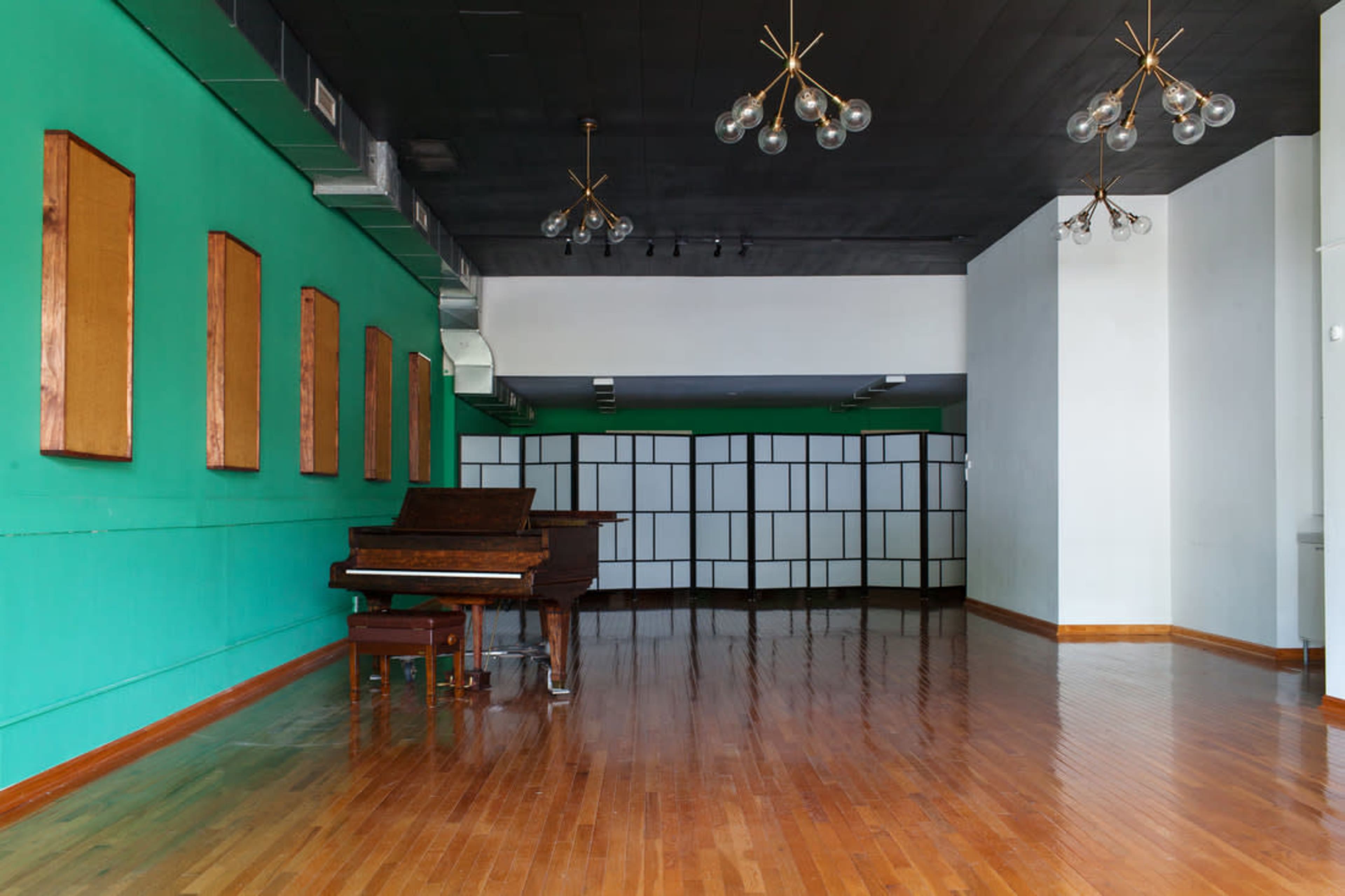 An empty room with polished wooden floors, a green accent wall with wooden panels, and a grand piano near the center, illuminated by modern light fixtures.
