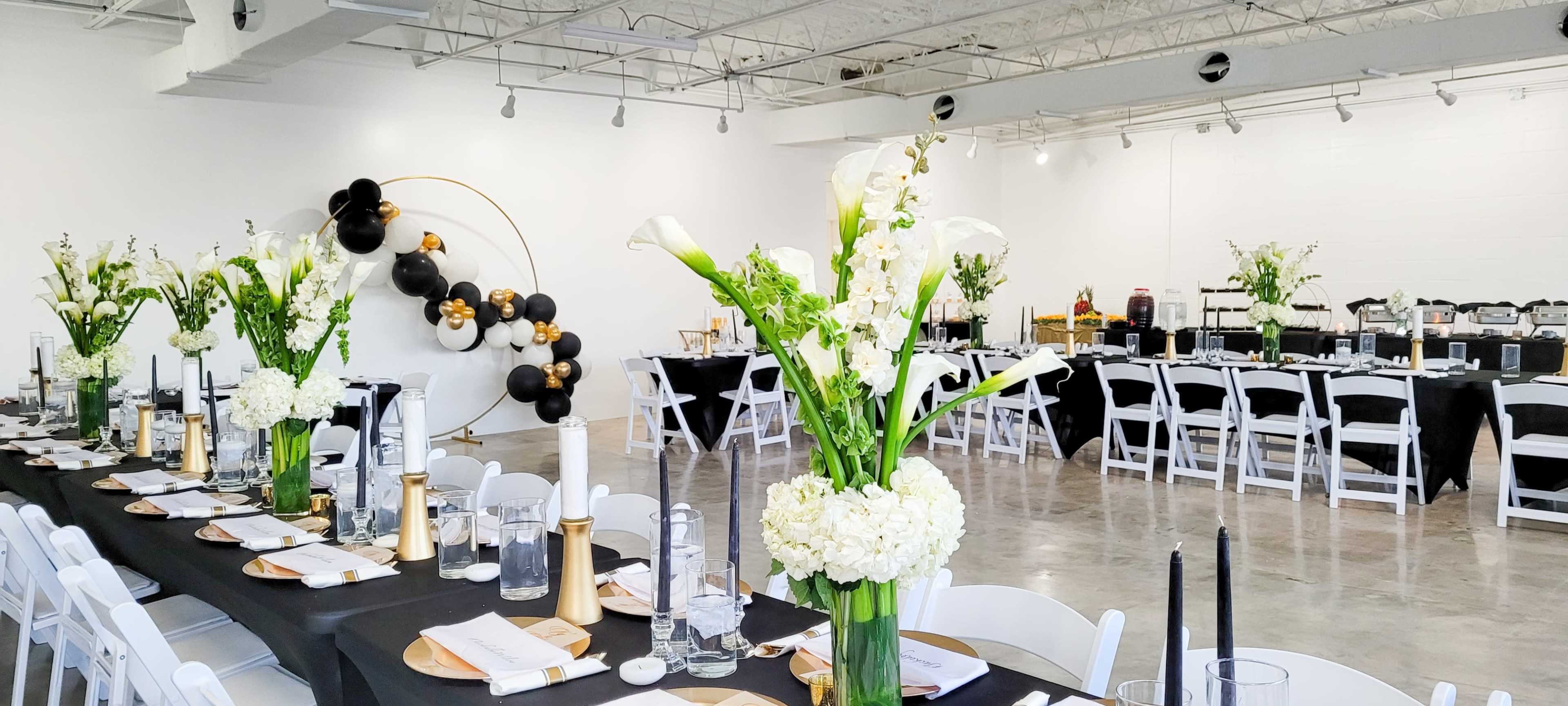 A banquet hall elegantly set up with long tables adorned with white flowers, black tablecloths, and gold accents, with a decorative arrangement of black and gold balloons in the background.