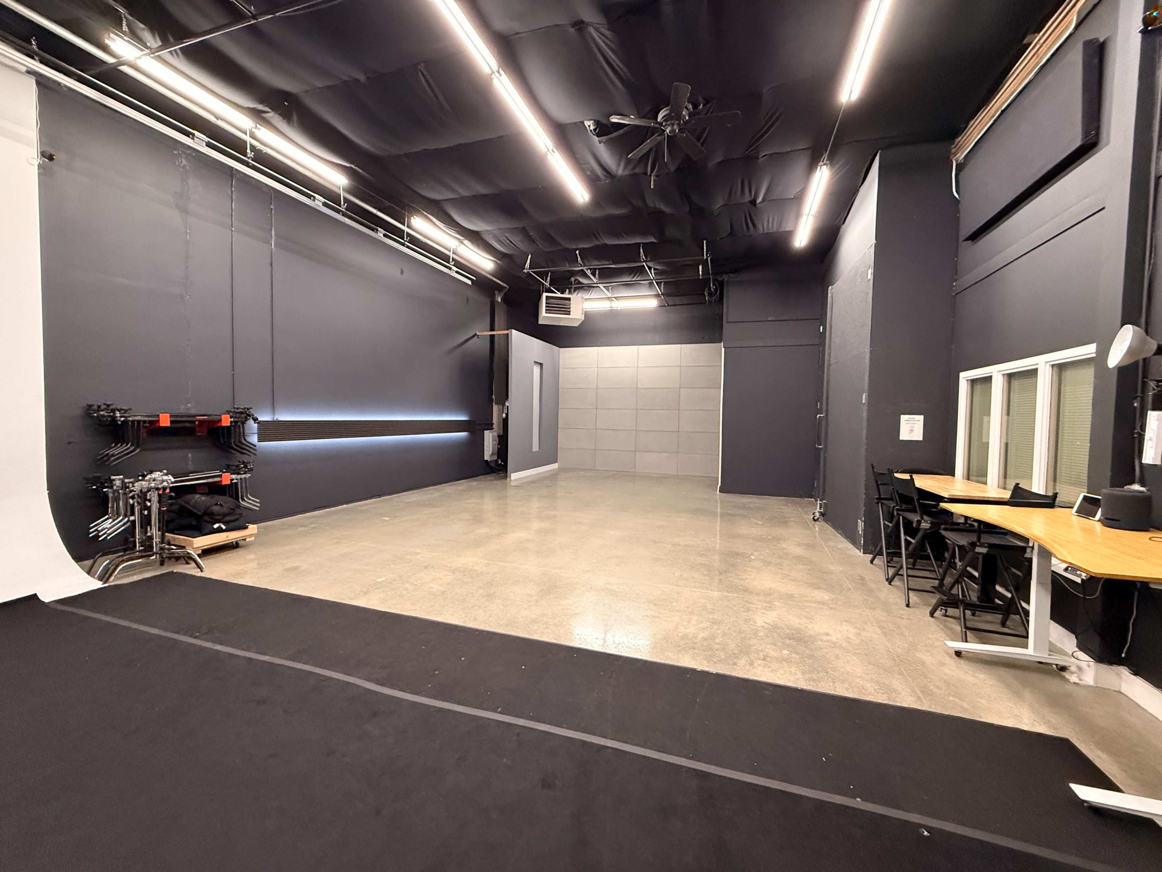 The image shows a spacious studio with gray walls, bright overhead lights, and a polished concrete floor, featuring a backdrop setup on one side and tables and chairs along the opposite wall.