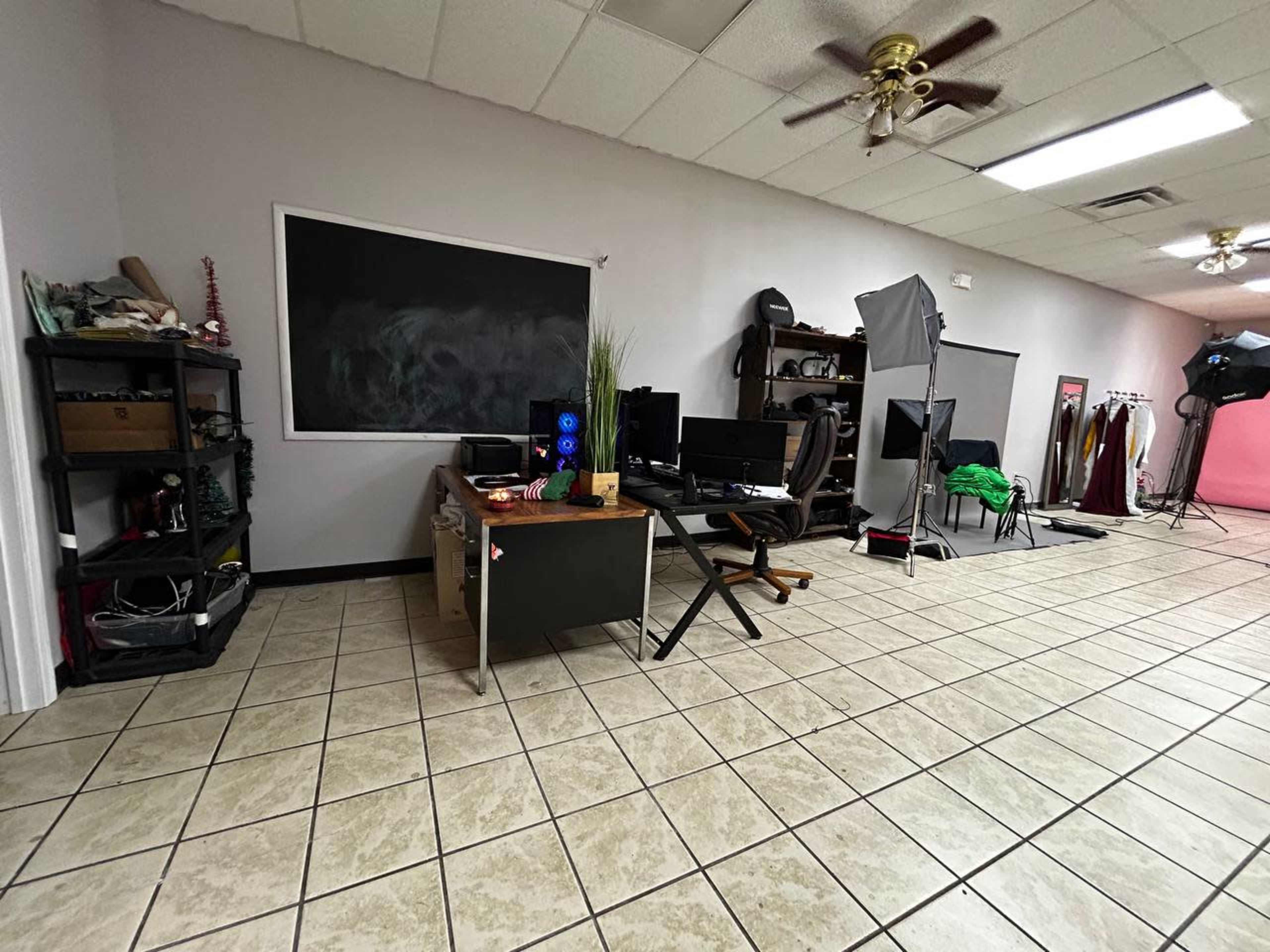 A spacious interior with tile flooring, featuring a desk with a computer setup, storage shelves, a chalkboard, and various photography equipment.