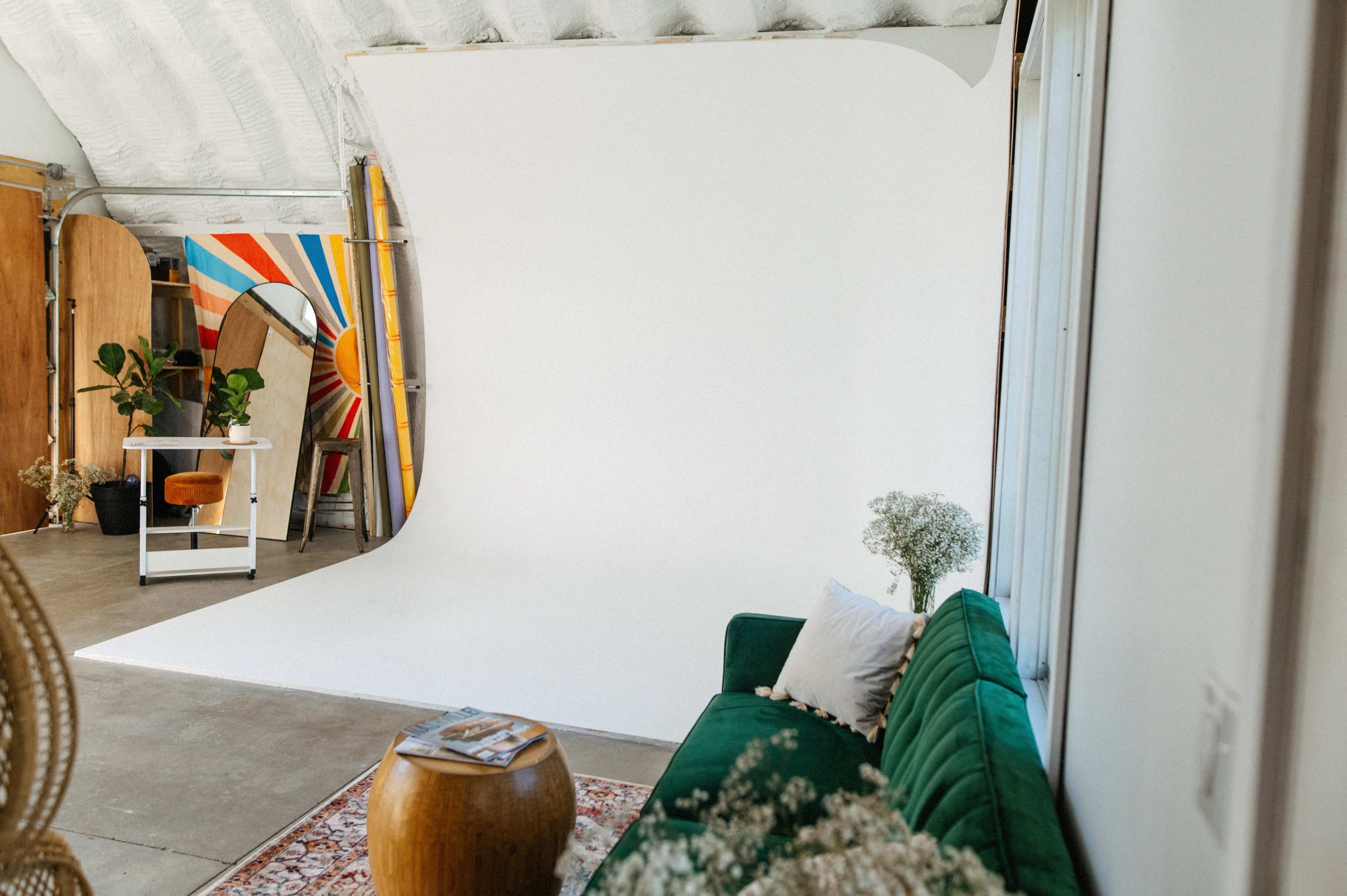 A bright photography studio with a white backdrop, a green couch, a round wooden table, and decorative elements including a chair and a full-length mirror.