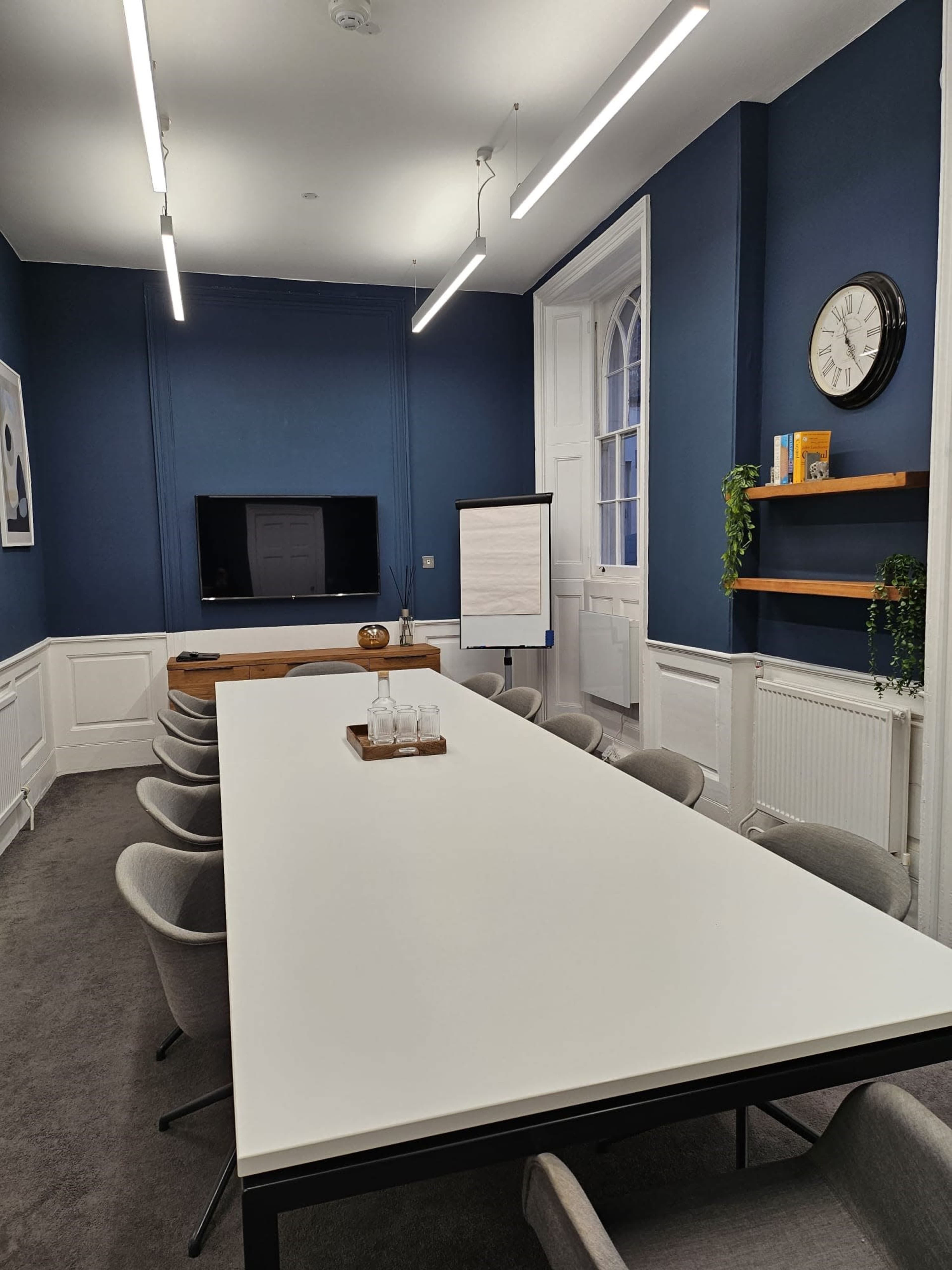 A modern conference room with a long white table, several grey chairs, a television on the wall, and decorative shelves.