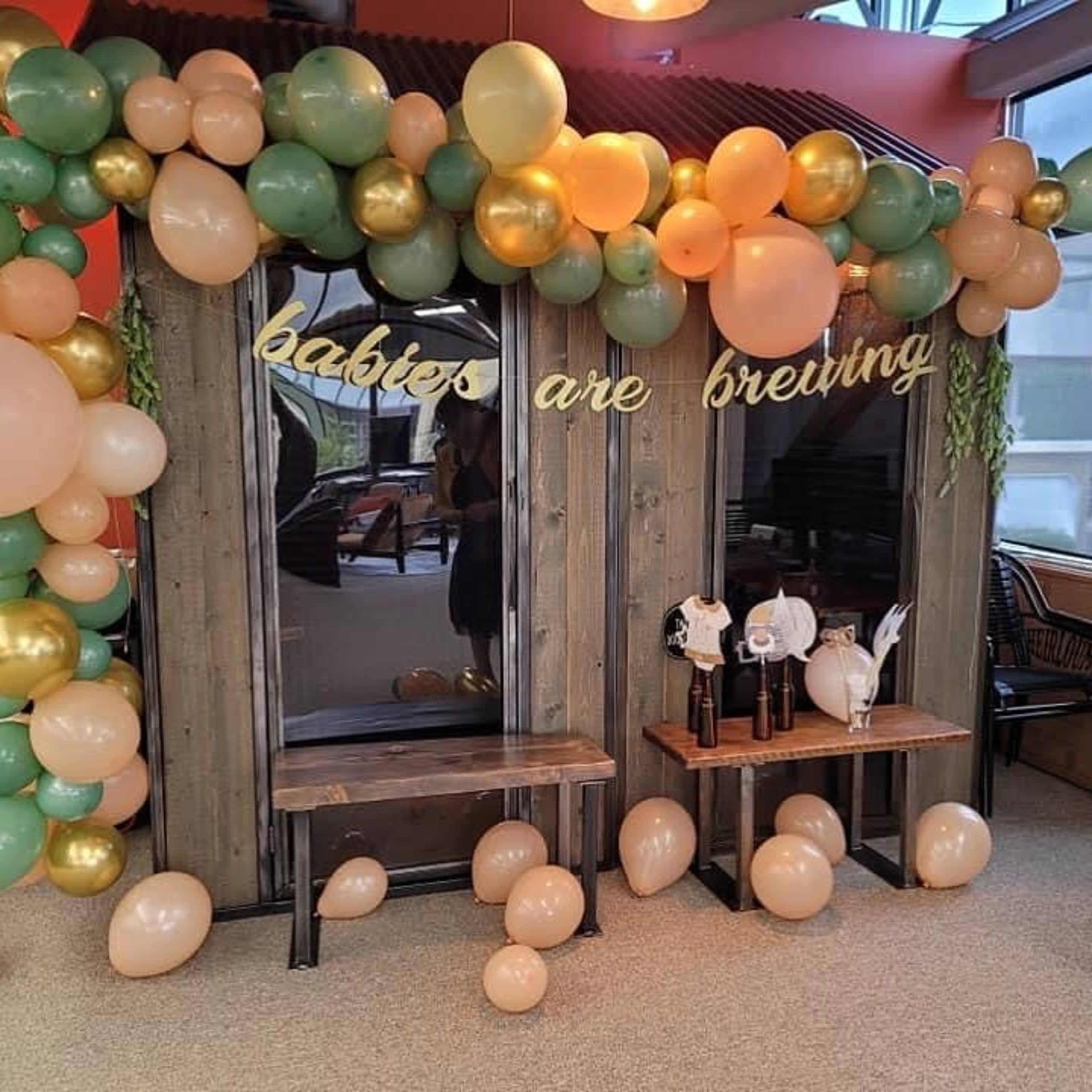 The image shows a decorated area with a wooden backdrop featuring the words "babies are brewing," surrounded by clusters of balloons in pastel pink, gold, and green, along with two small tables displaying baby-themed items.
