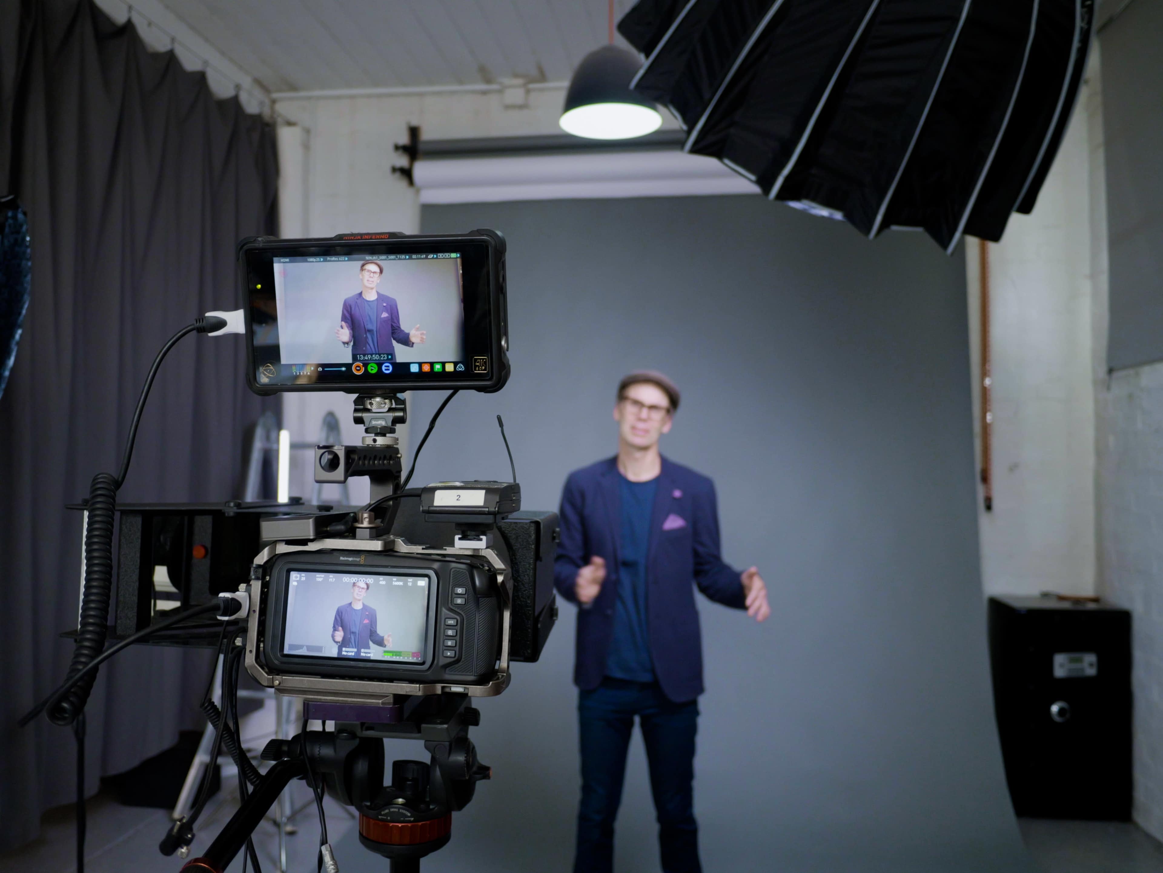A man is standing in front of a gray backdrop while being filmed by a camera set up with monitors and lighting equipment.