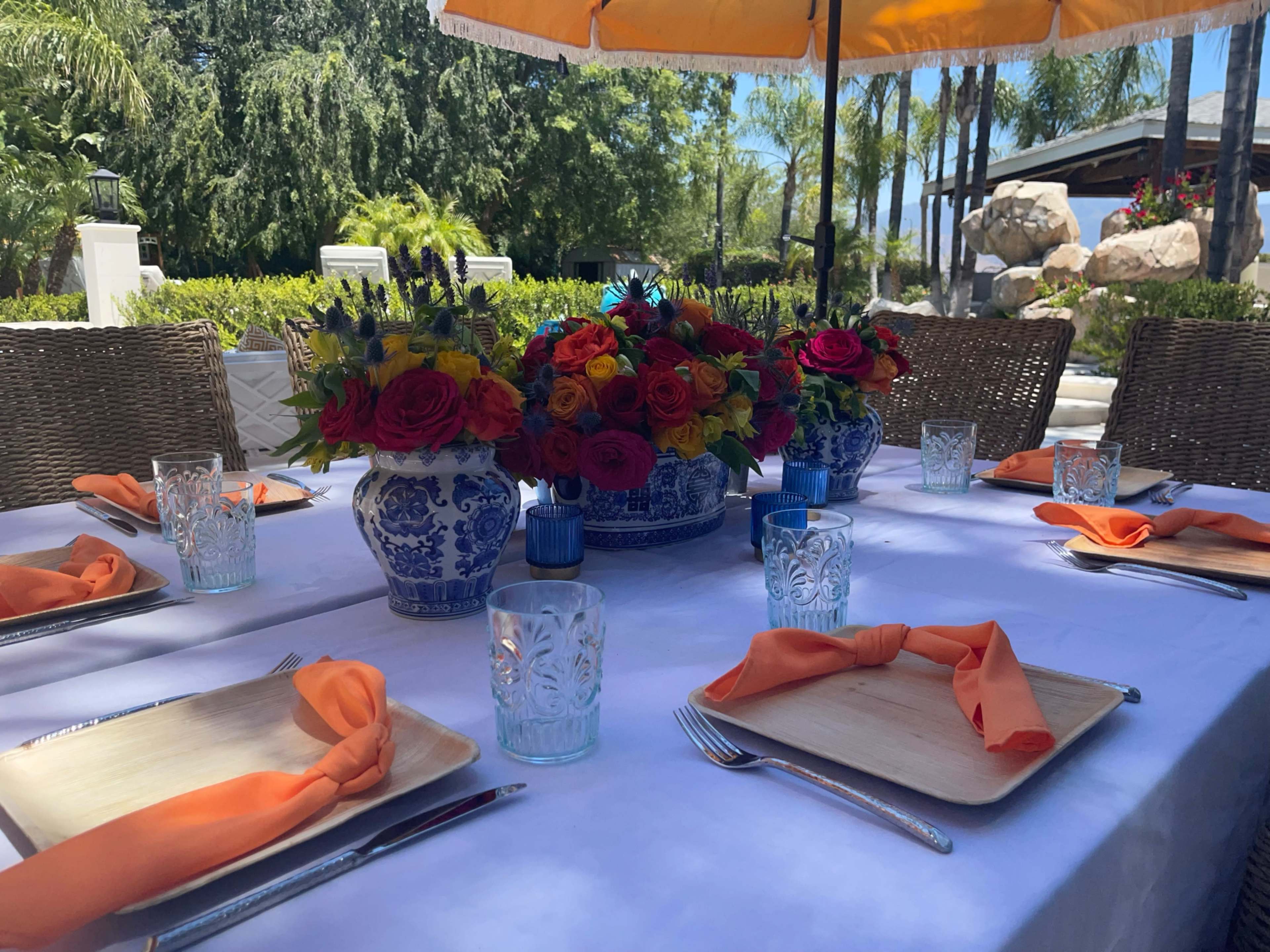 A dining table is set outdoors with a colorful floral centerpiece, orange napkins, and glassware, surrounded by greenery and palm trees.