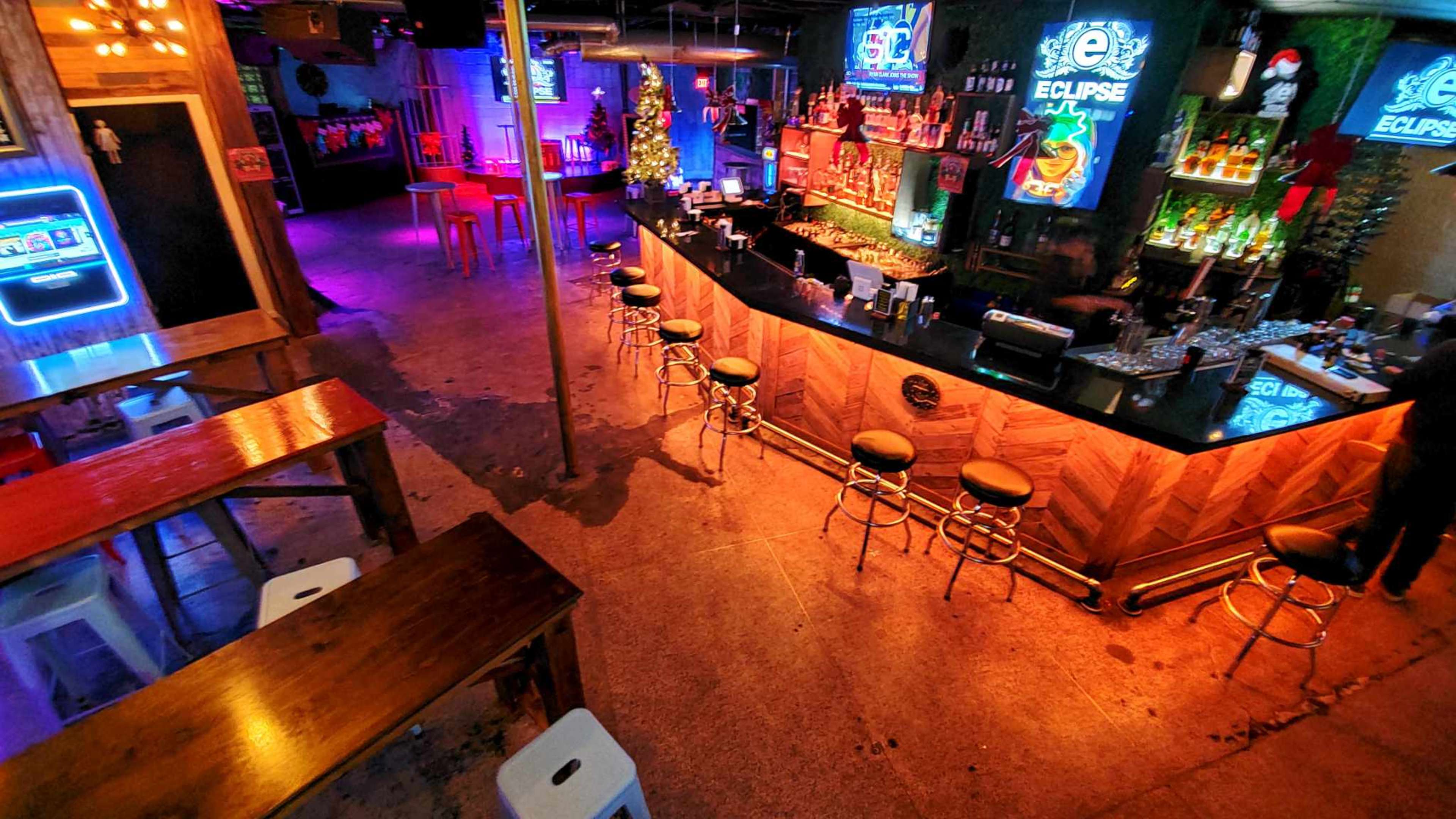 The image shows a bar area with a long countertop, several bar stools, colorful neon lights, and shelves stocked with various liquor bottles.