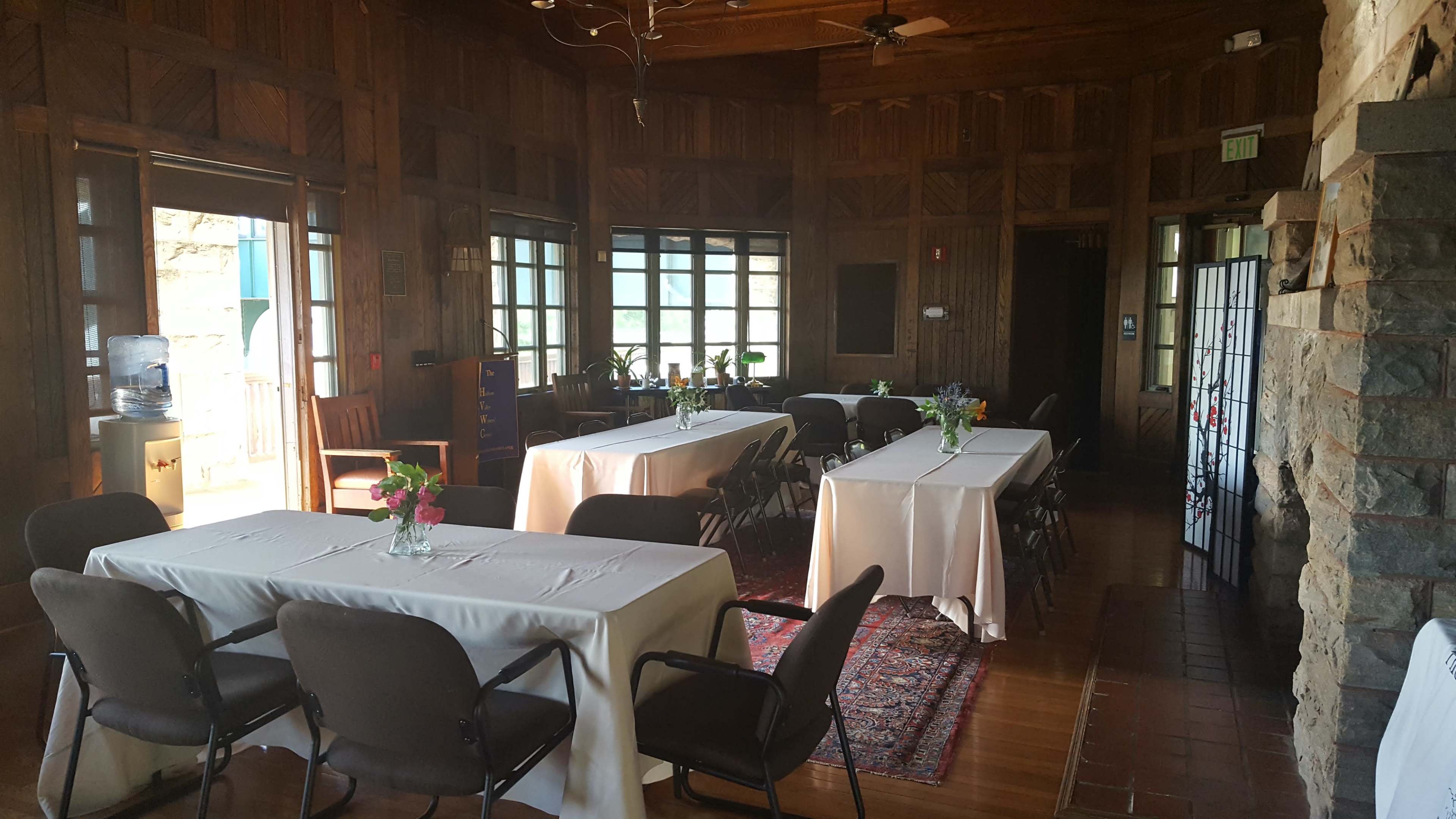 The image shows a wooden-paneled room set up with several long tables covered in white tablecloths, accompanied by chairs and flower centerpieces.