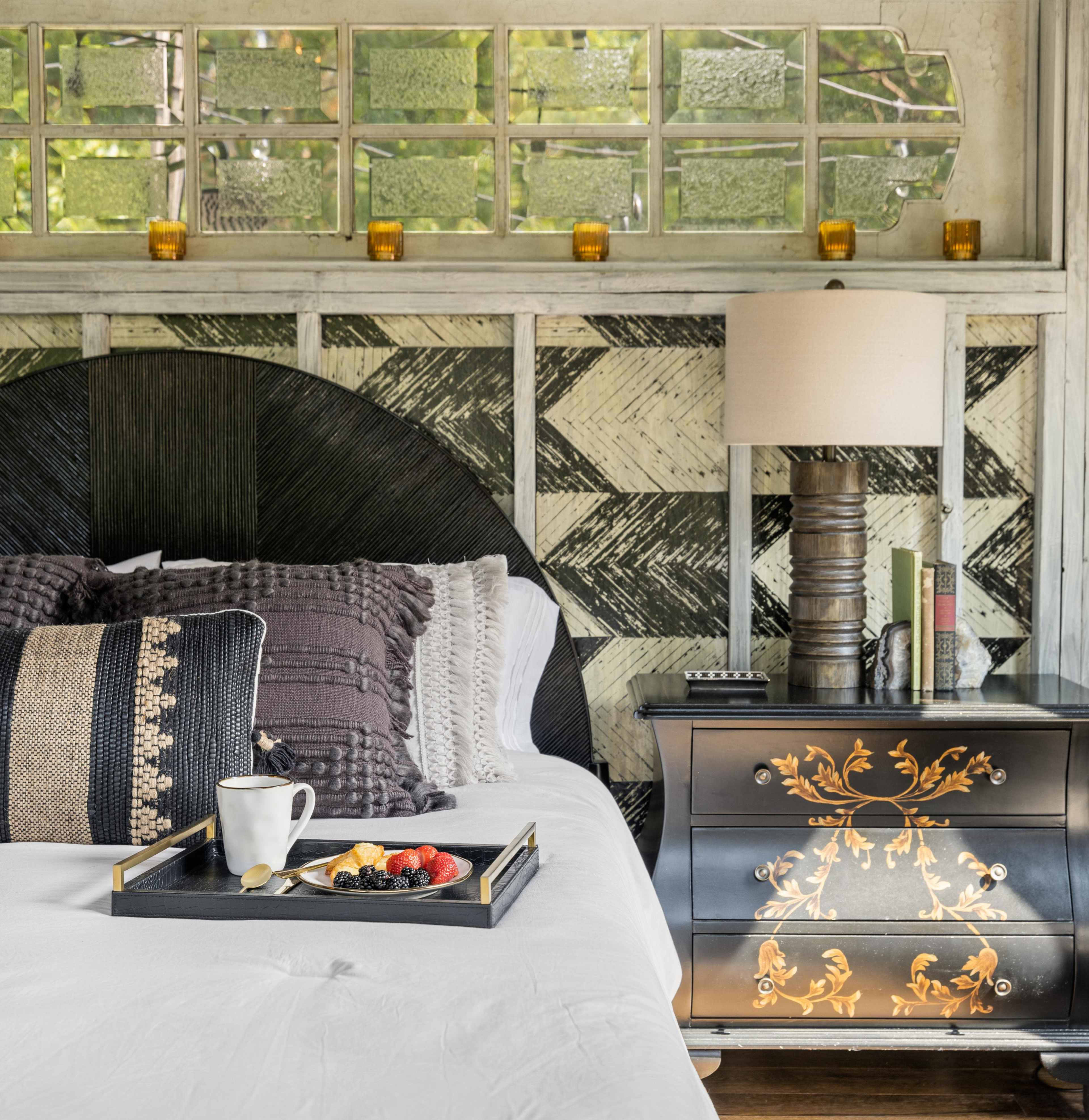 A neatly made bed with a textured headboard, accompanied by a nightstand holding a decorative lamp and a tray featuring fruit and a mug.