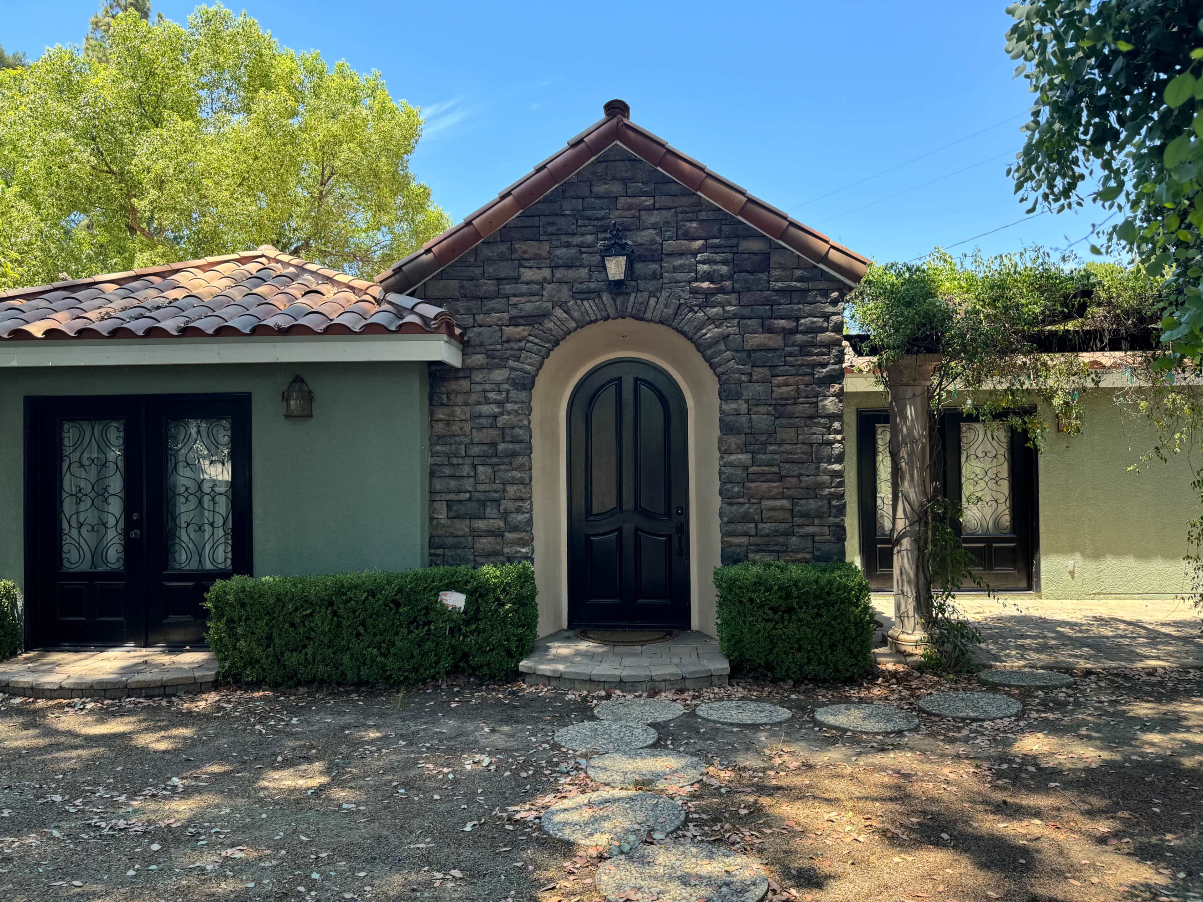 A stone and stucco house features a clay tile roof, a large arched entryway, and well-maintained landscaping with shrubs and stone pathways.