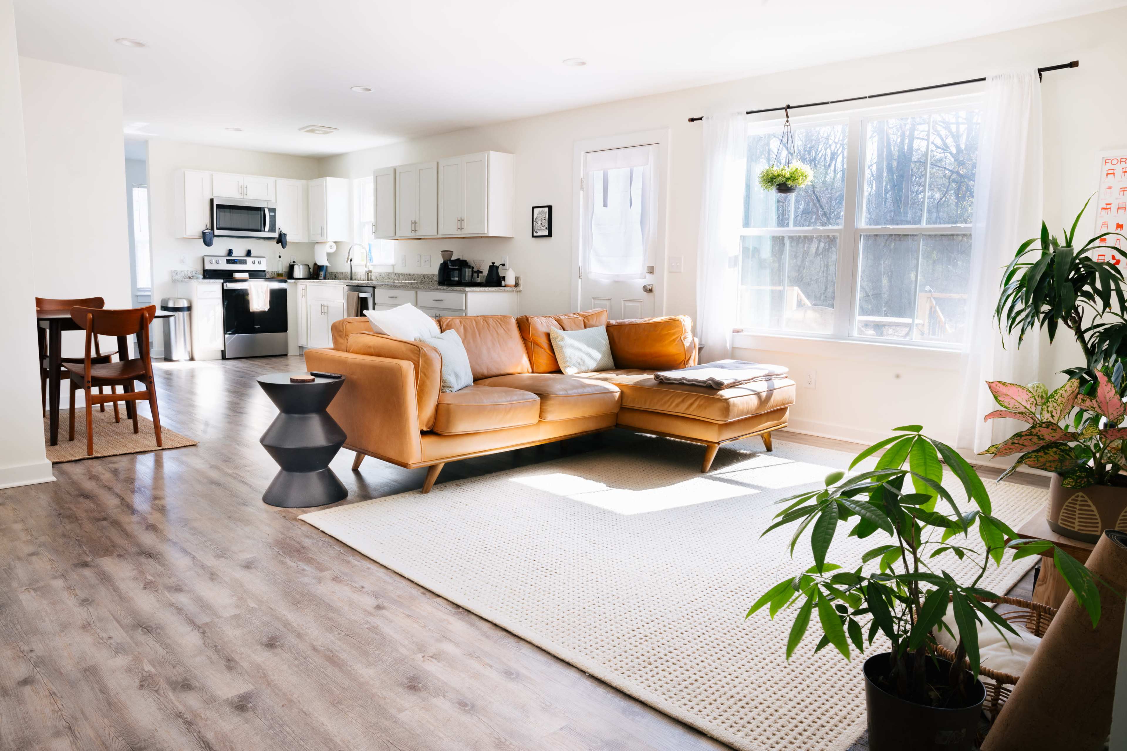 The image shows a bright, modern living room with a tan leather sofa, a round side table, a woven area rug, and plants, leading into a minimalist kitchen area.