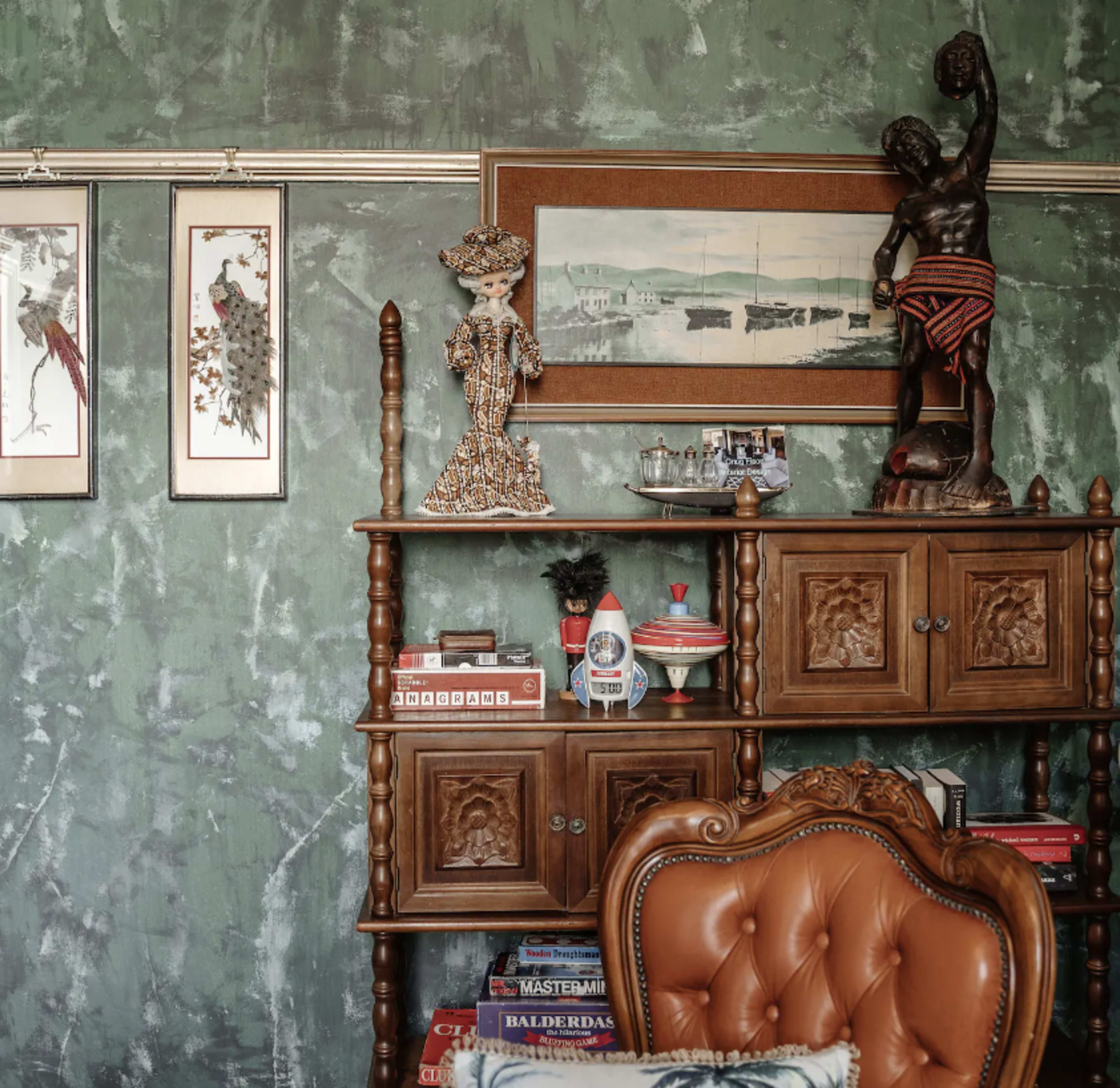 The image shows a wooden shelf adorned with decorative items, framed artwork, and a vintage chair against a textured green wall.