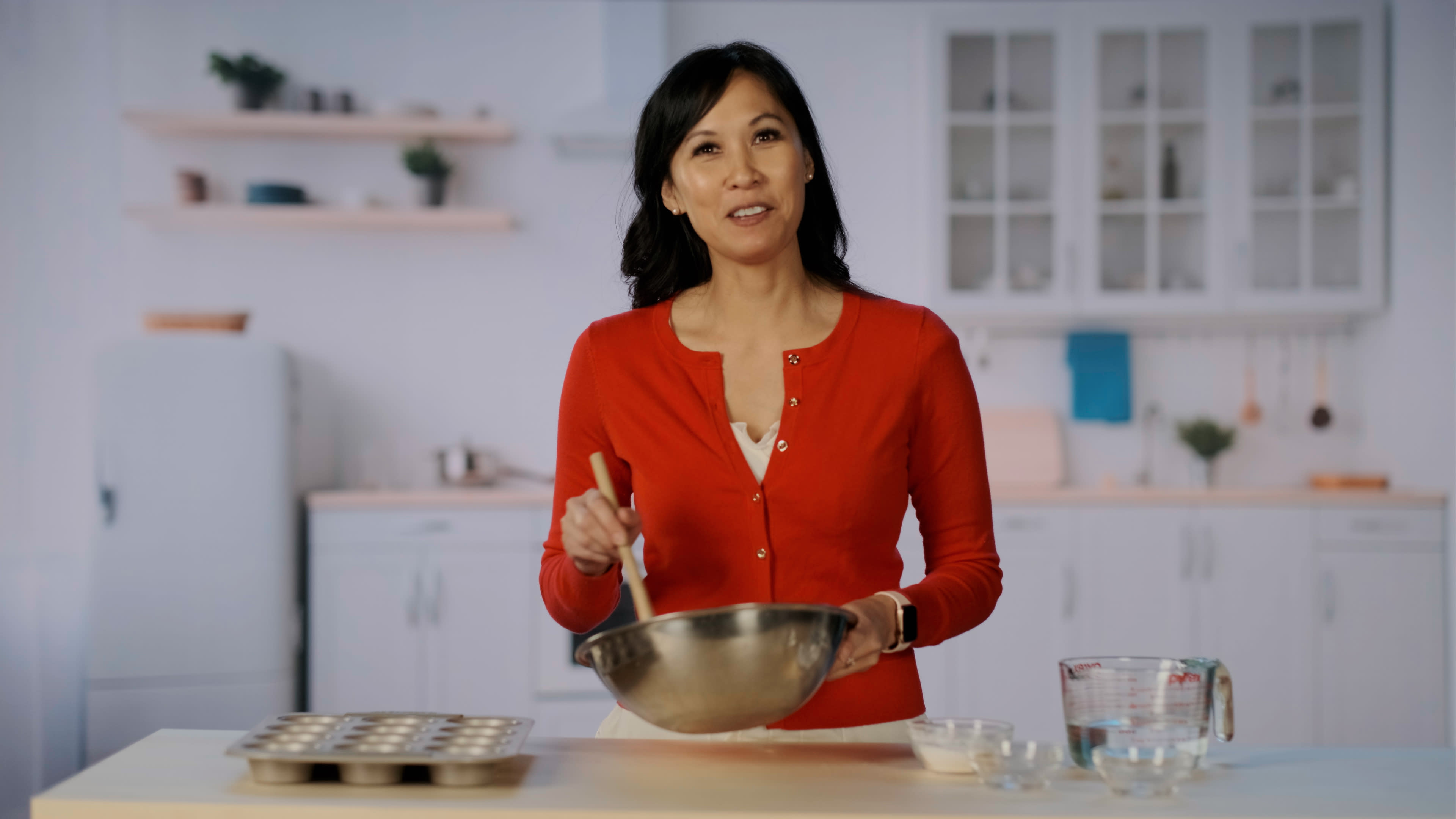 A woman in a red cardigan is stirring a mixture in a bowl while standing at a kitchen counter.