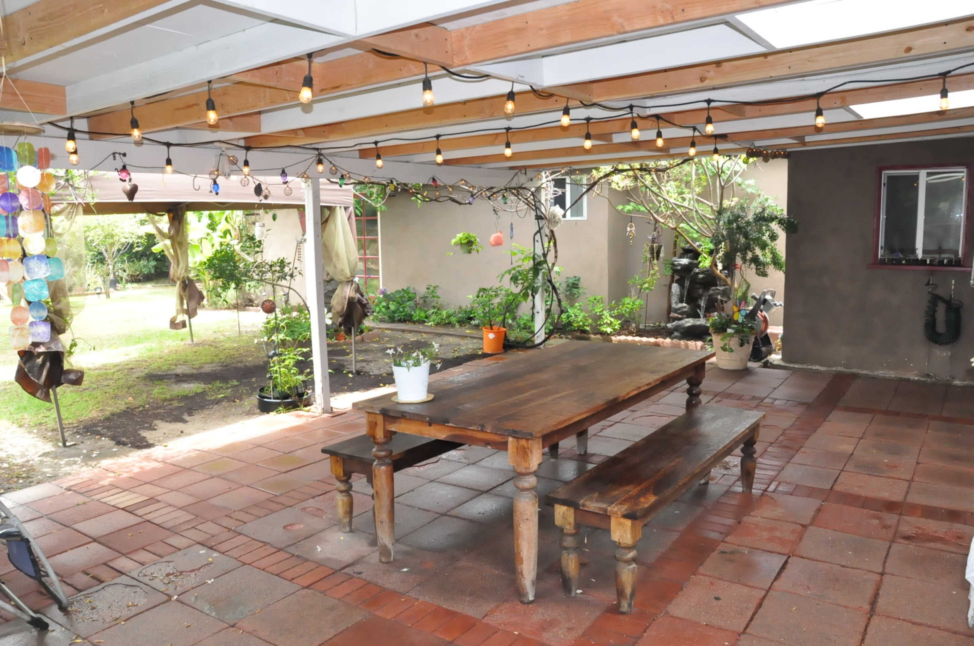 A covered outdoor patio area featuring a wooden table and benches, with string lights overhead and green plants in the background.