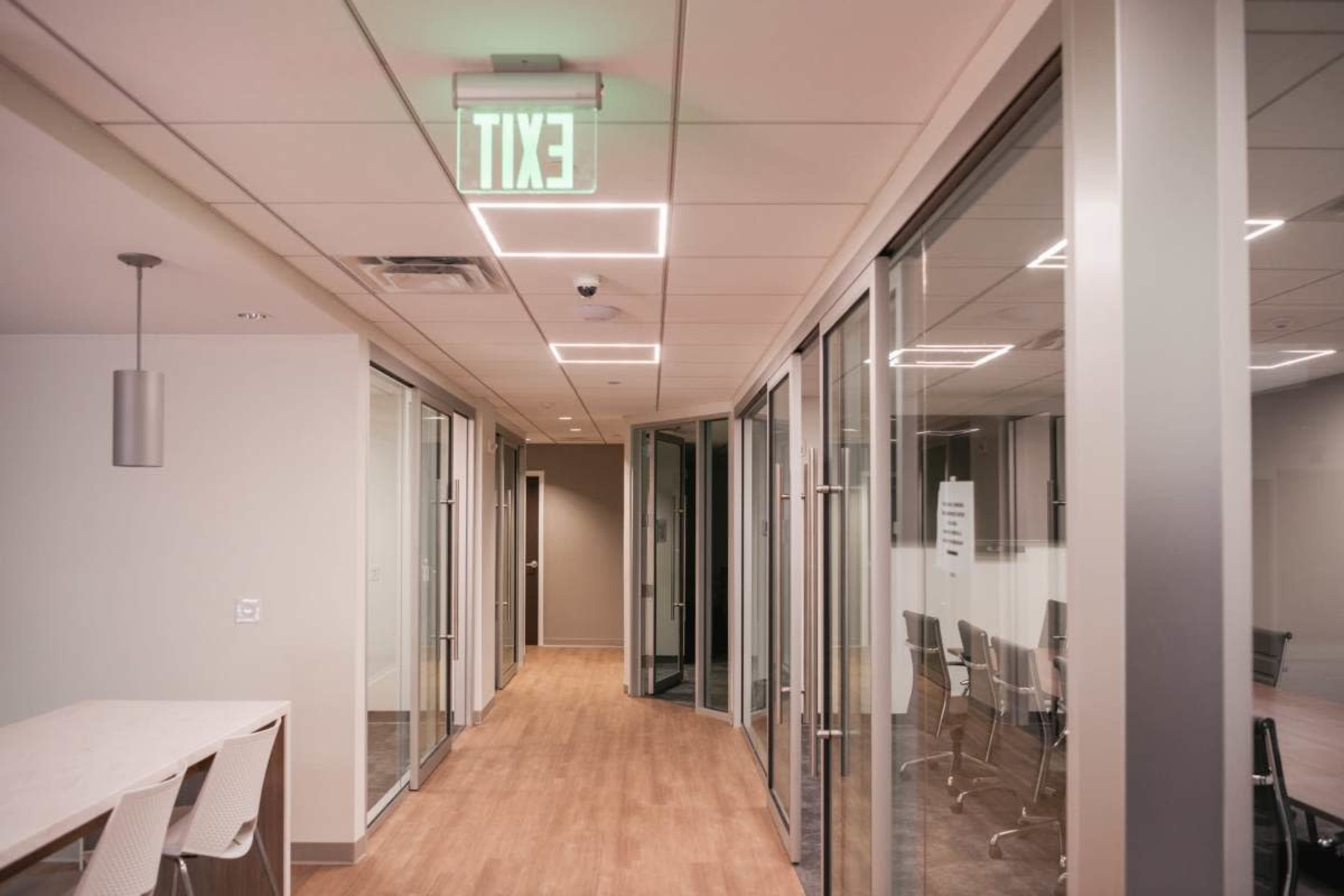 The image shows a modern office corridor with glass-walled meeting rooms and an illuminated exit sign hanging from the ceiling.