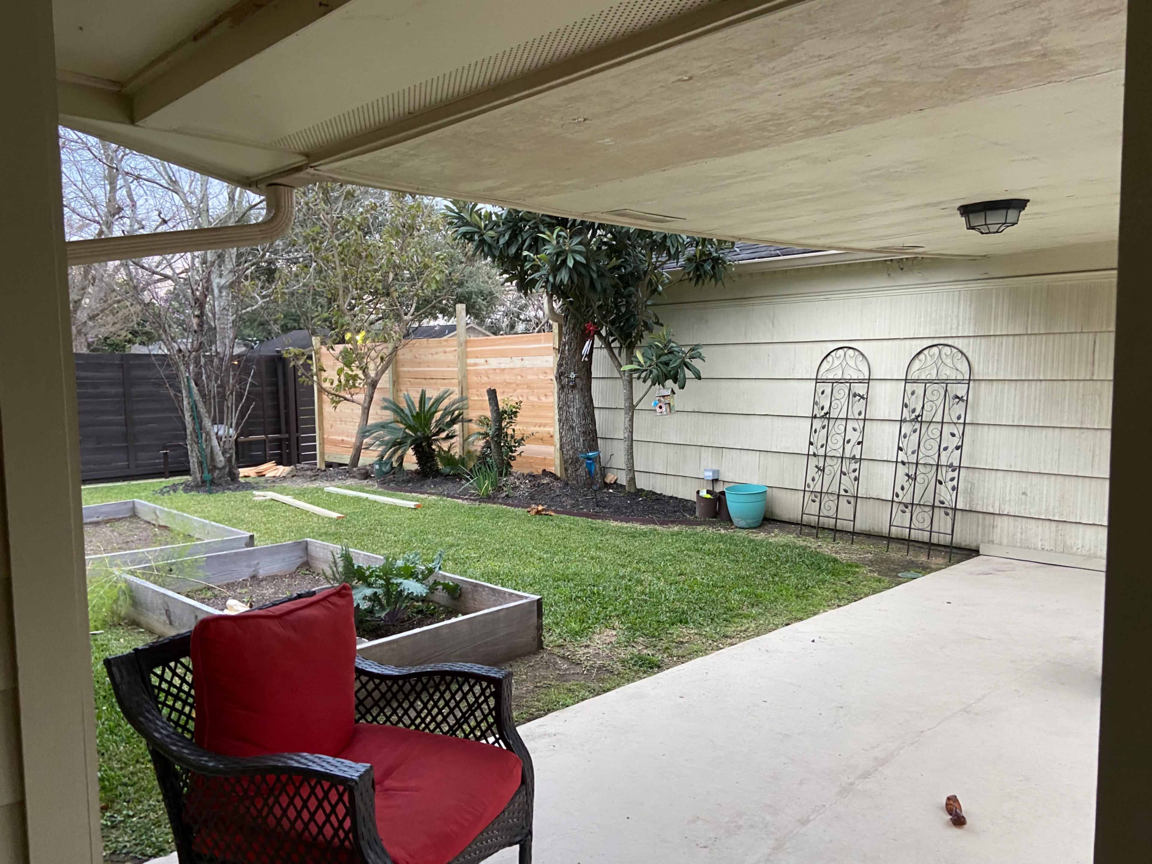 A patio area with a red cushioned chair overlooks a grassy yard with raised garden beds and a wooden fence in the background.