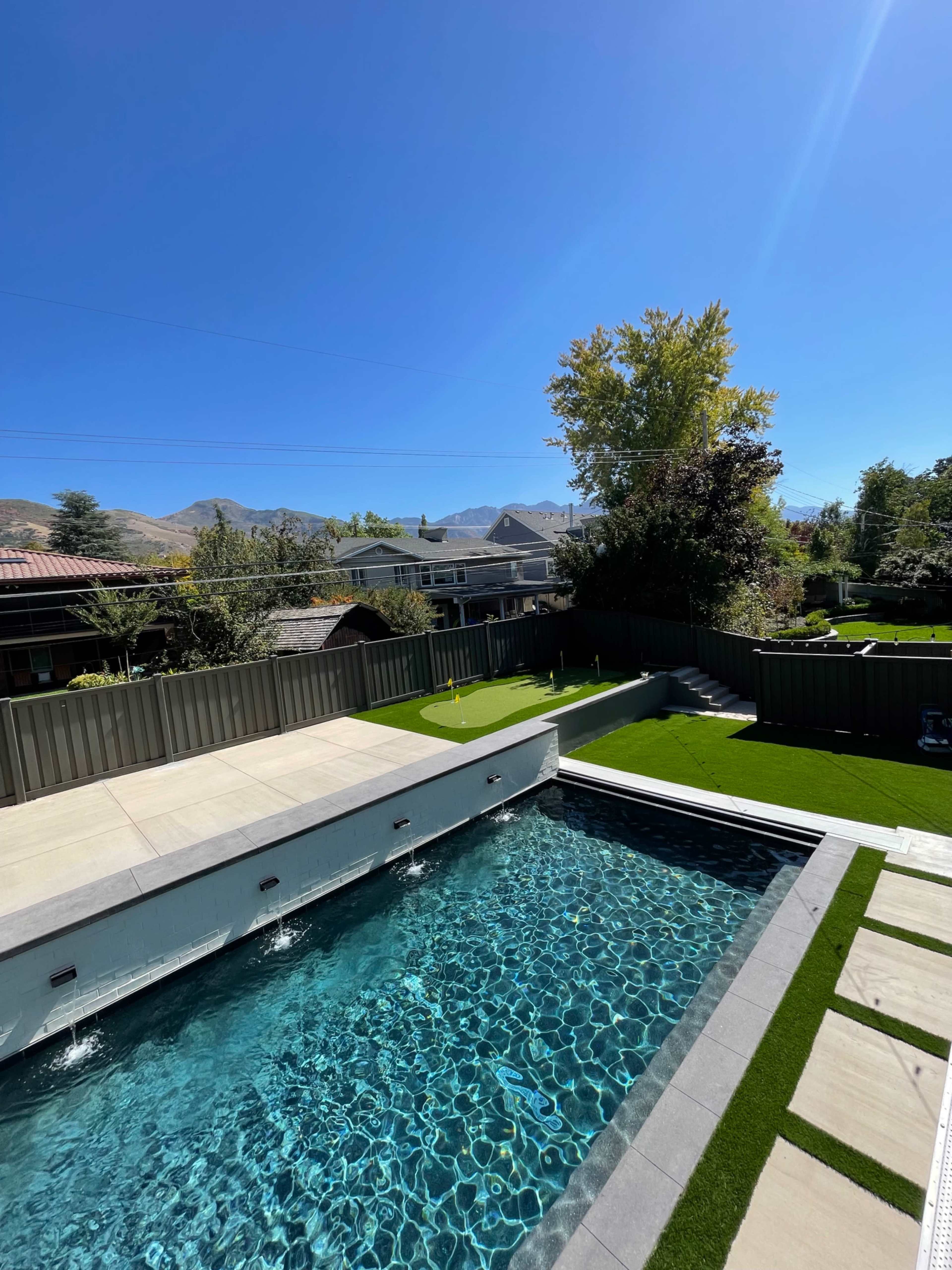 A clear blue pool with stone decking is surrounded by green grass and homes, framed by distant mountains under a bright sky.