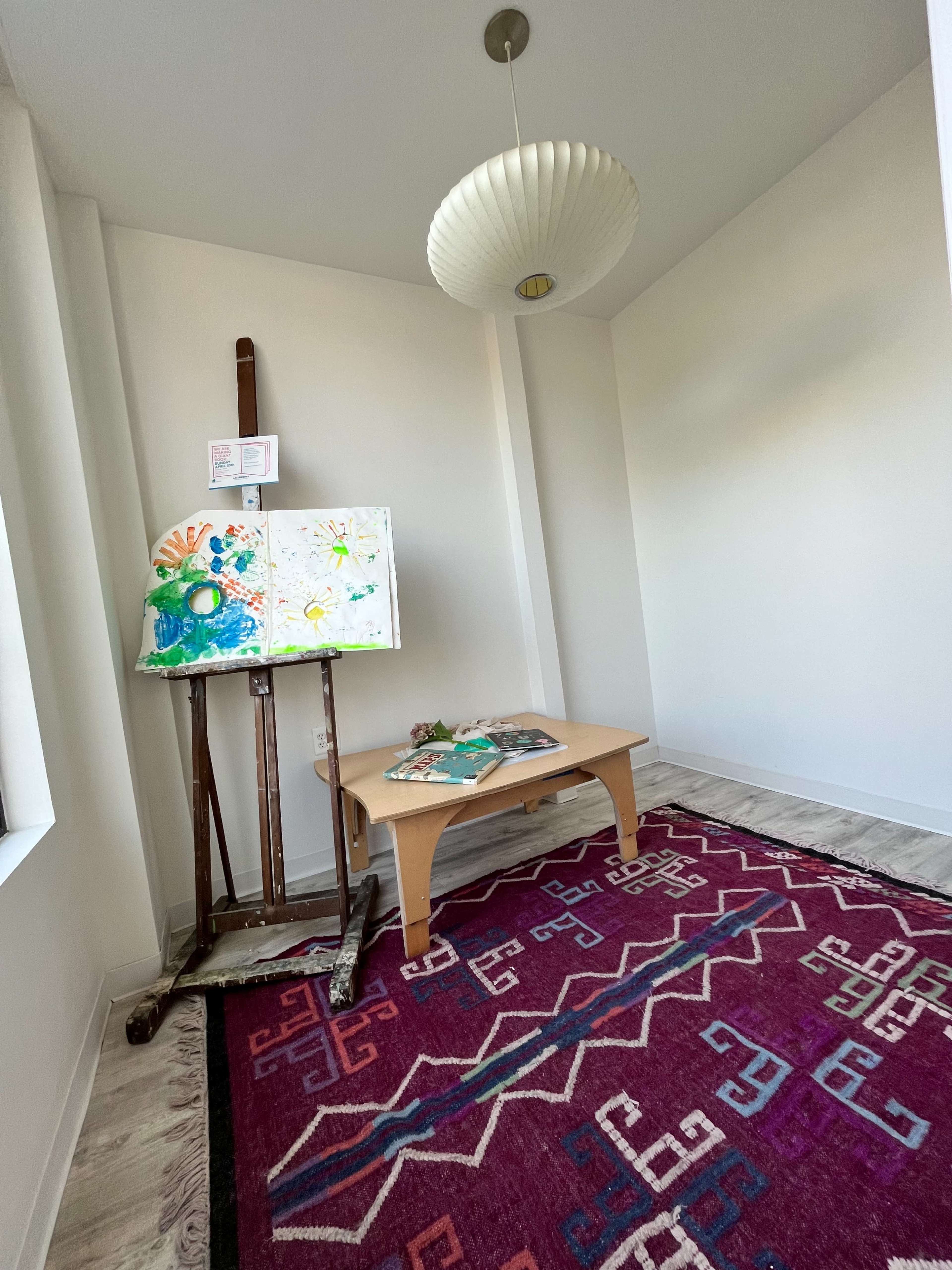A well-lit corner of a room with an easel displaying colorful artwork, a small wooden table, and a patterned rug on the floor.