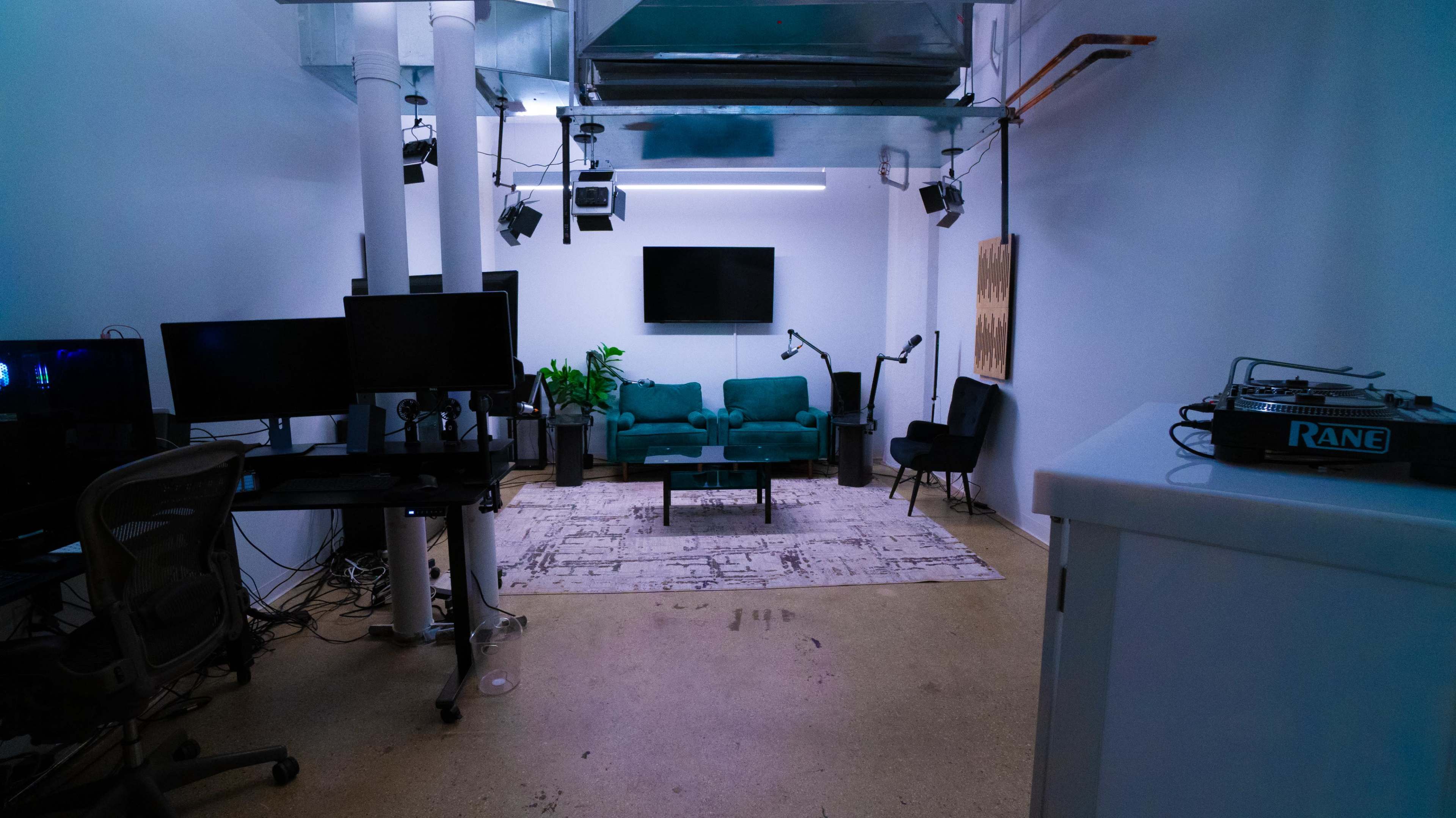 The image shows a modern, sparsely furnished office space with a teal couch, a large wall-mounted TV, and several computer monitors along with a DJ setup on a side table.