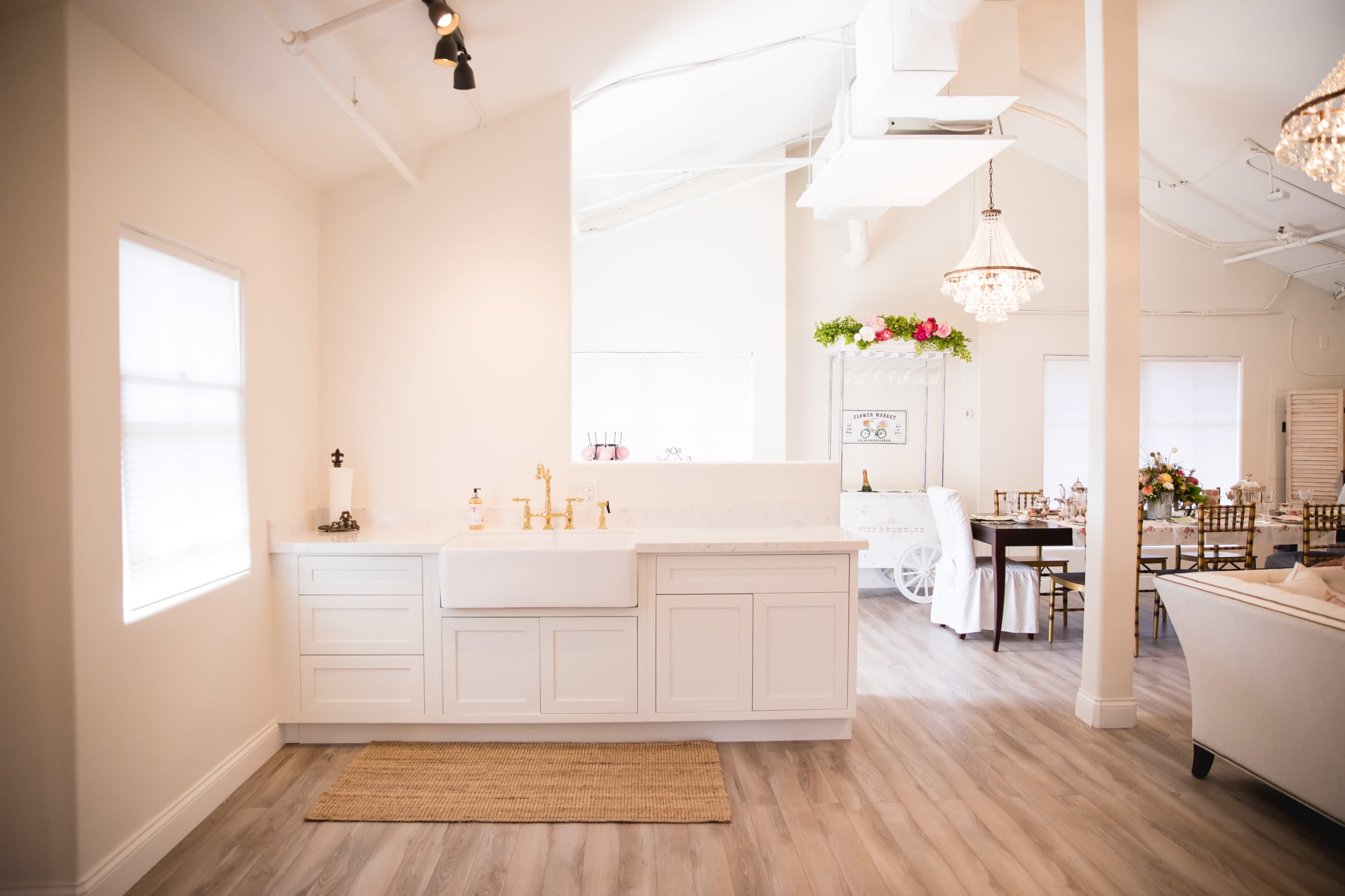 A modern kitchen area features a farmhouse sink, white cabinetry, and a view into a dining space adorned with floral decorations and elegant lighting.