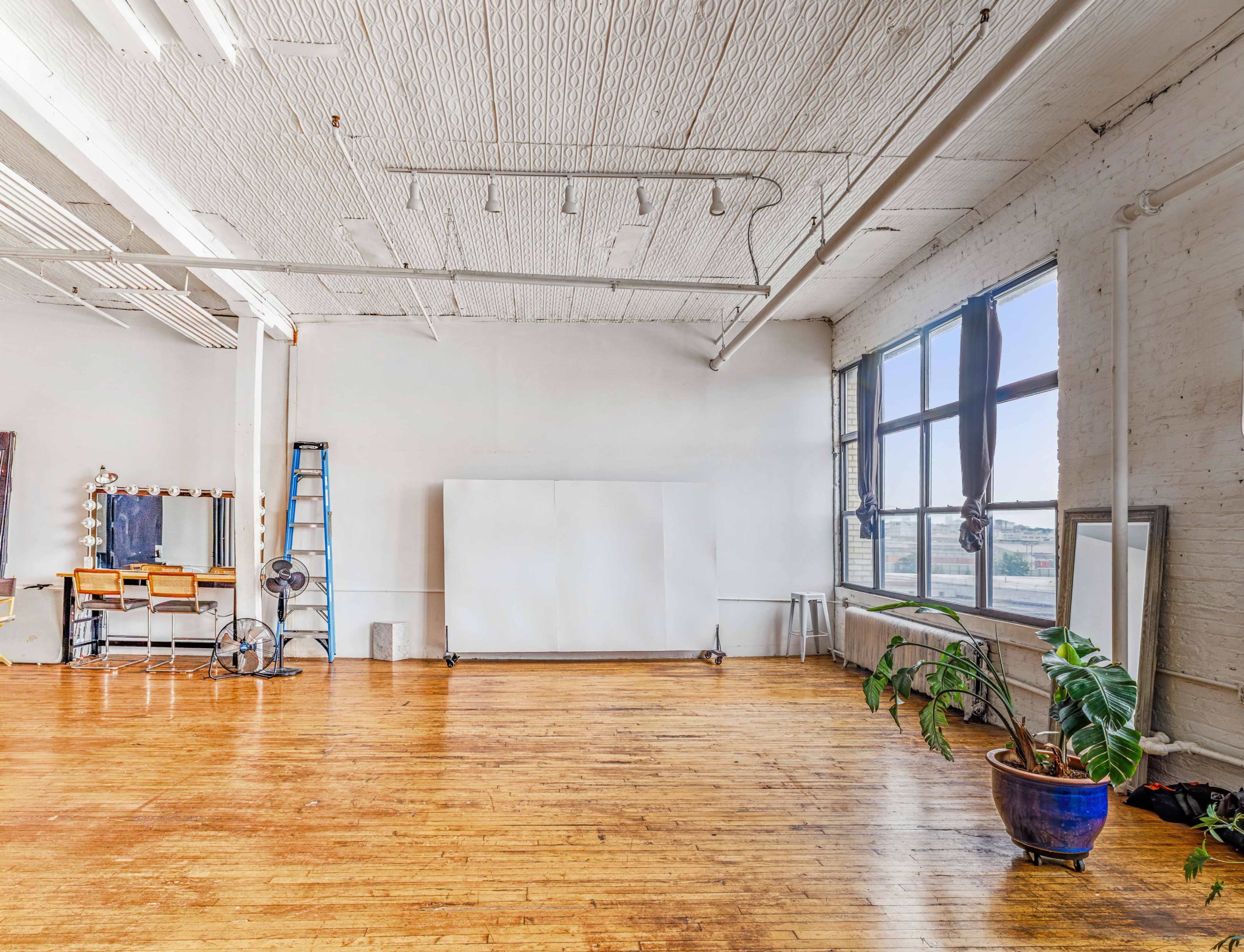 The image depicts a spacious, well-lit studio with wooden floors, large windows, and a blank white wall, along with a makeup station, a ladder, and a potted plant.