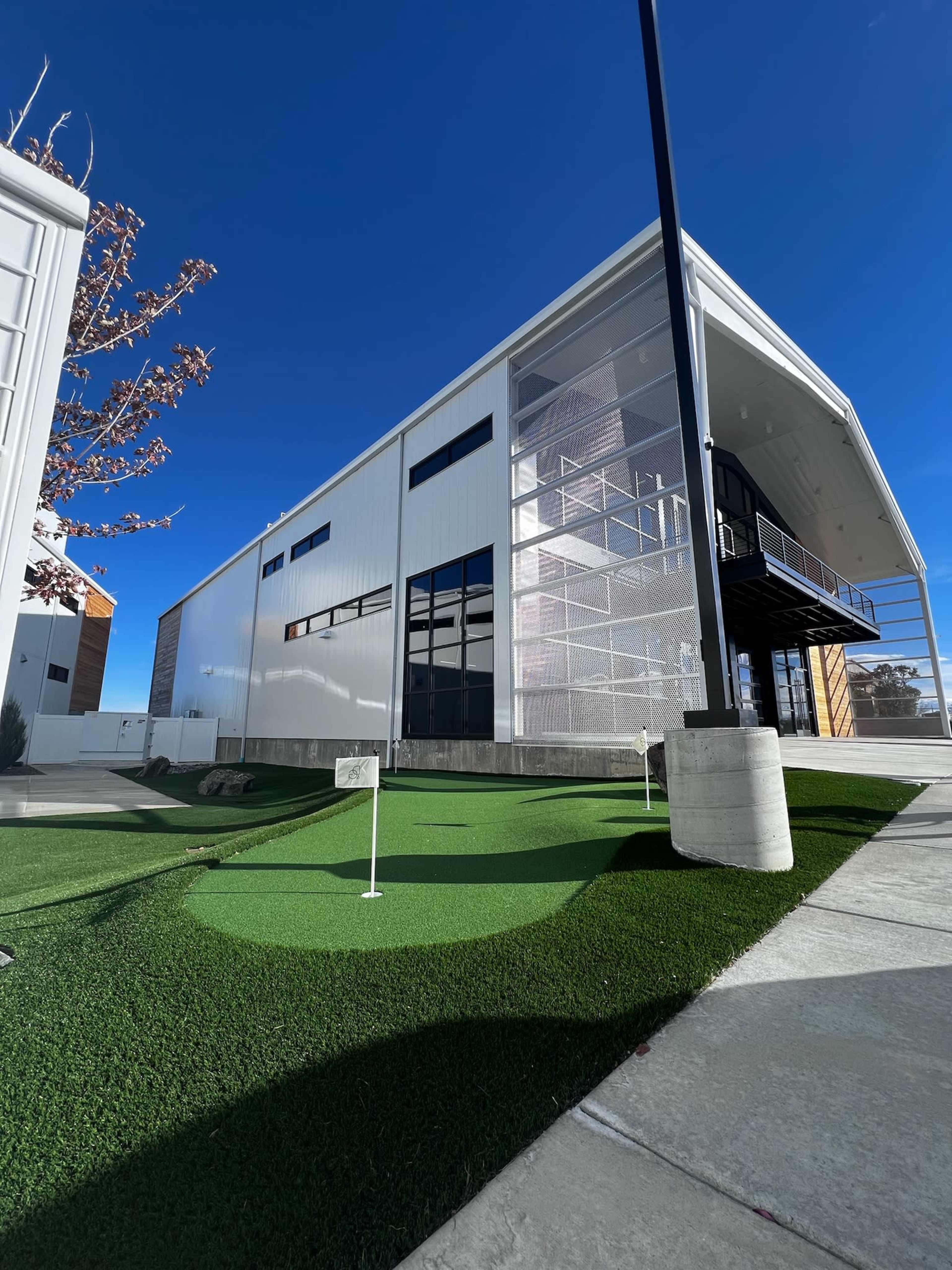 A modern building with a large glass front and a small artificial grass putting green in the foreground under a clear blue sky.