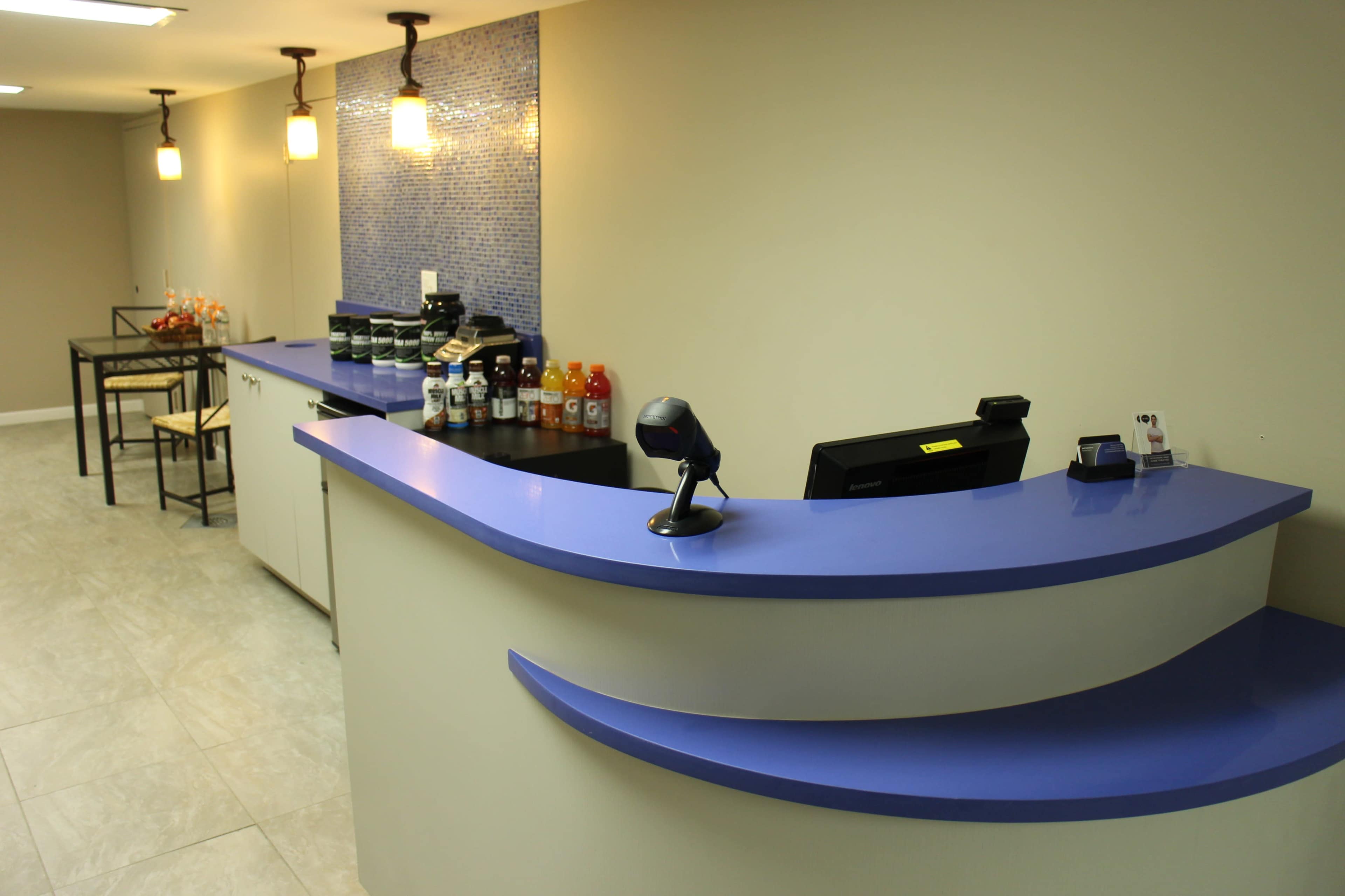 The image shows a modern reception desk with a curved blue countertop, accompanied by shelves of beverages and a seating area in a well-lit interior space.
