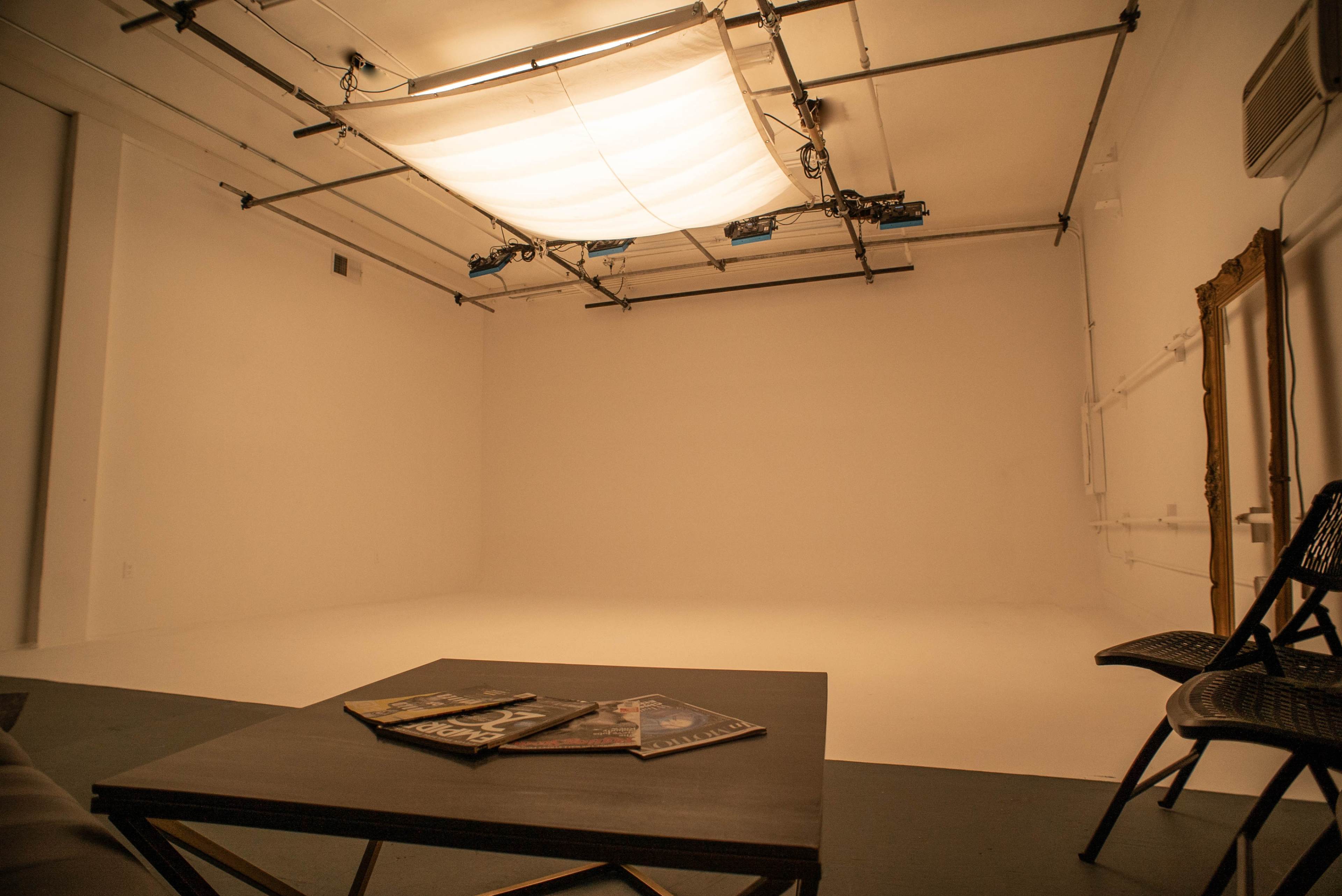 An empty photography studio with a softbox lighting setup, a small table with magazines, and two chairs against a wall.
