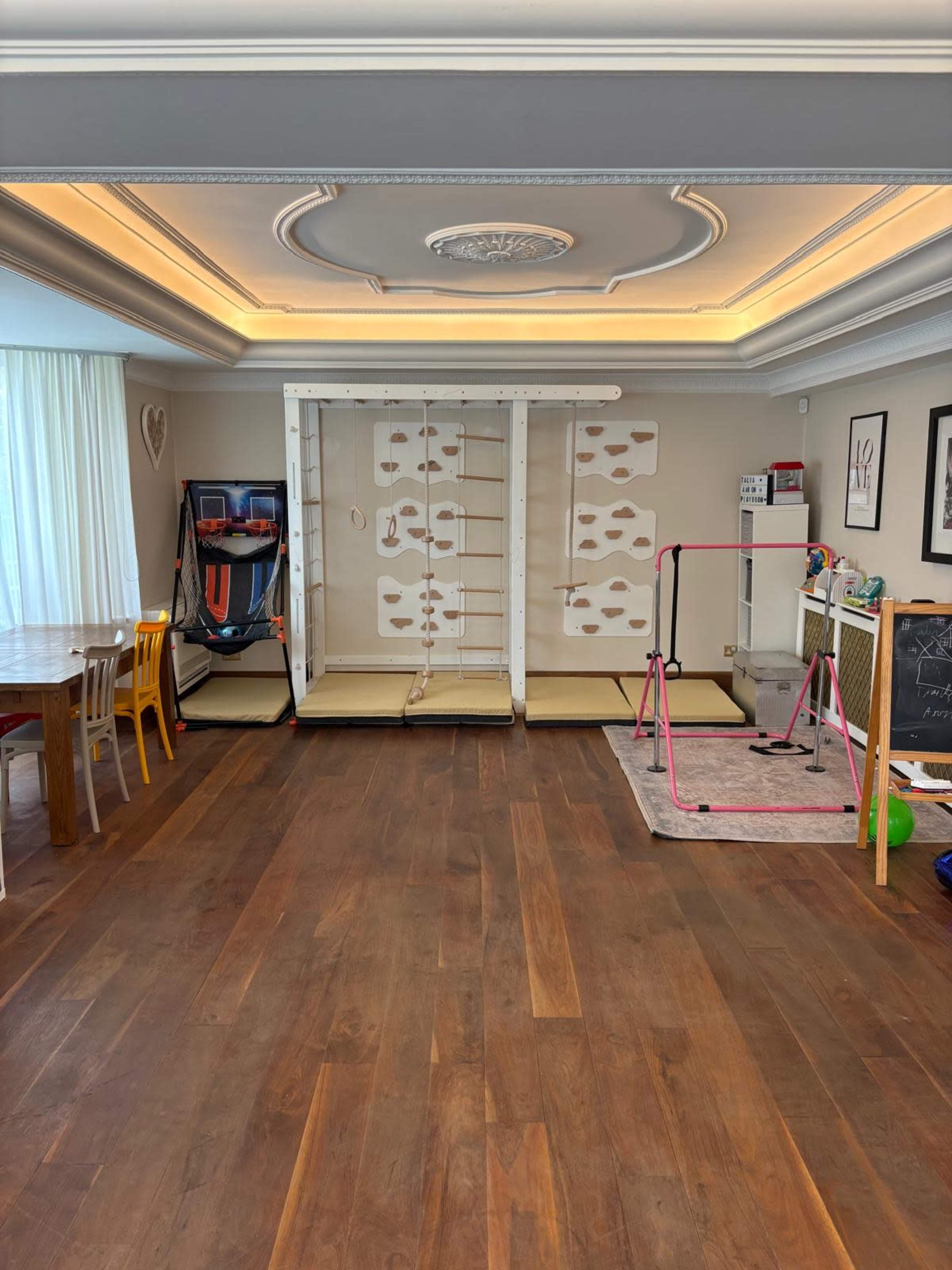 The image shows a spacious room with wooden flooring, featuring a climbing wall and play equipment on one side, a dining table and chairs on the other, and large windows allowing natural light in.