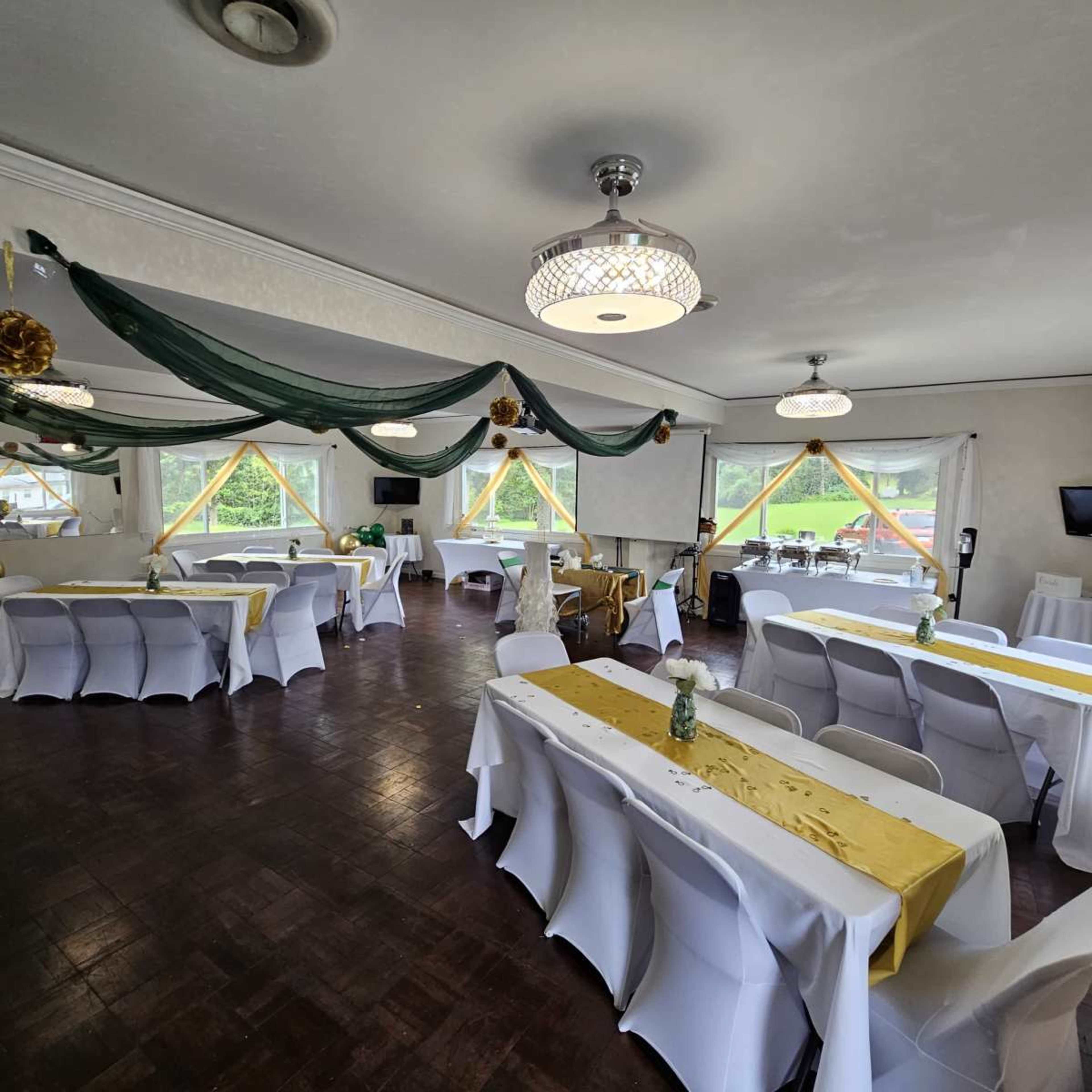 The image shows a well-arranged indoor event space featuring rows of tables covered with white linens and a gold accent, surrounded by large windows with decorative drapery.