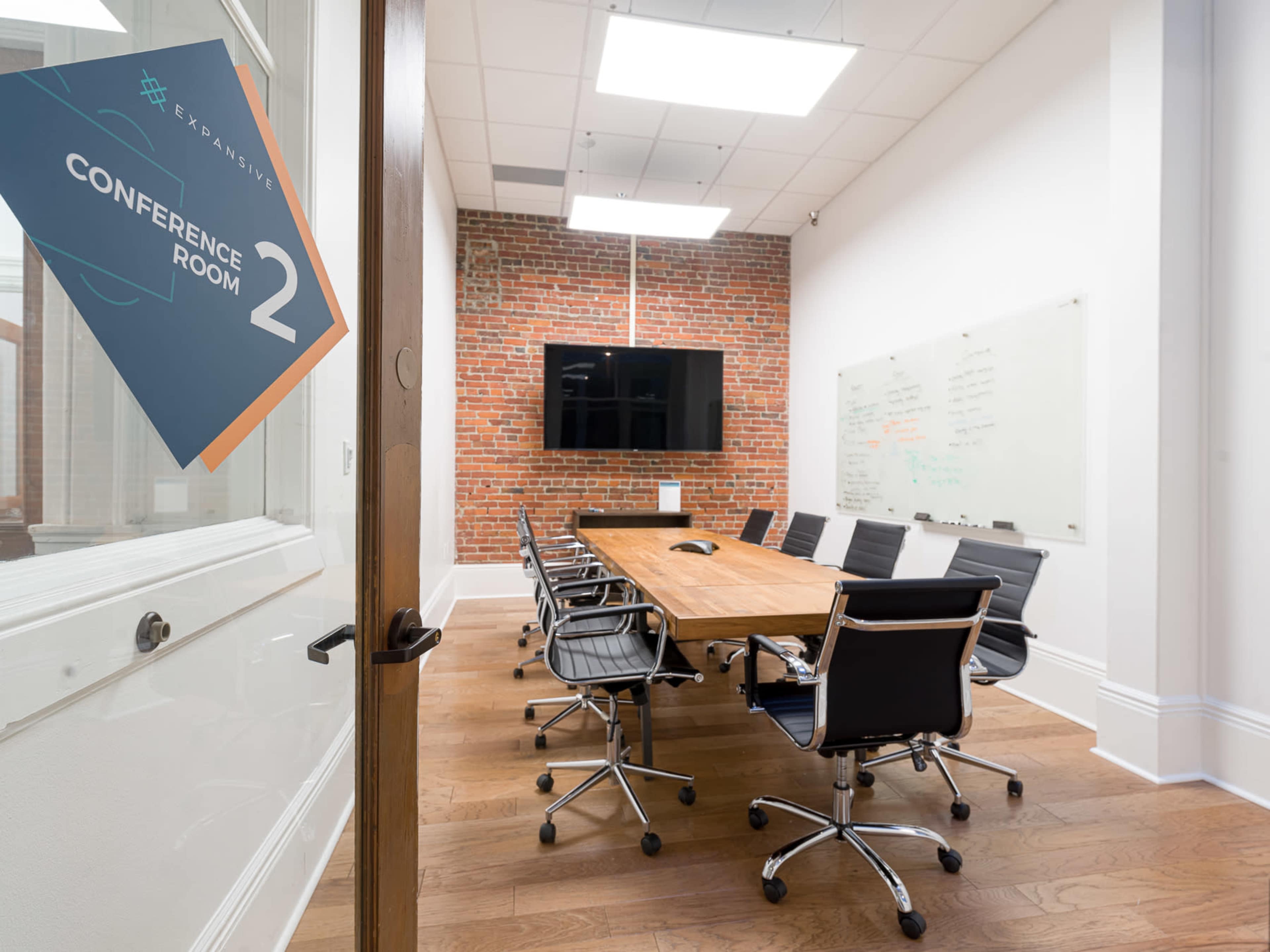A conference room featuring a large wooden table, several black chairs, a television screen on a brick wall, and a whiteboard for presentations.
