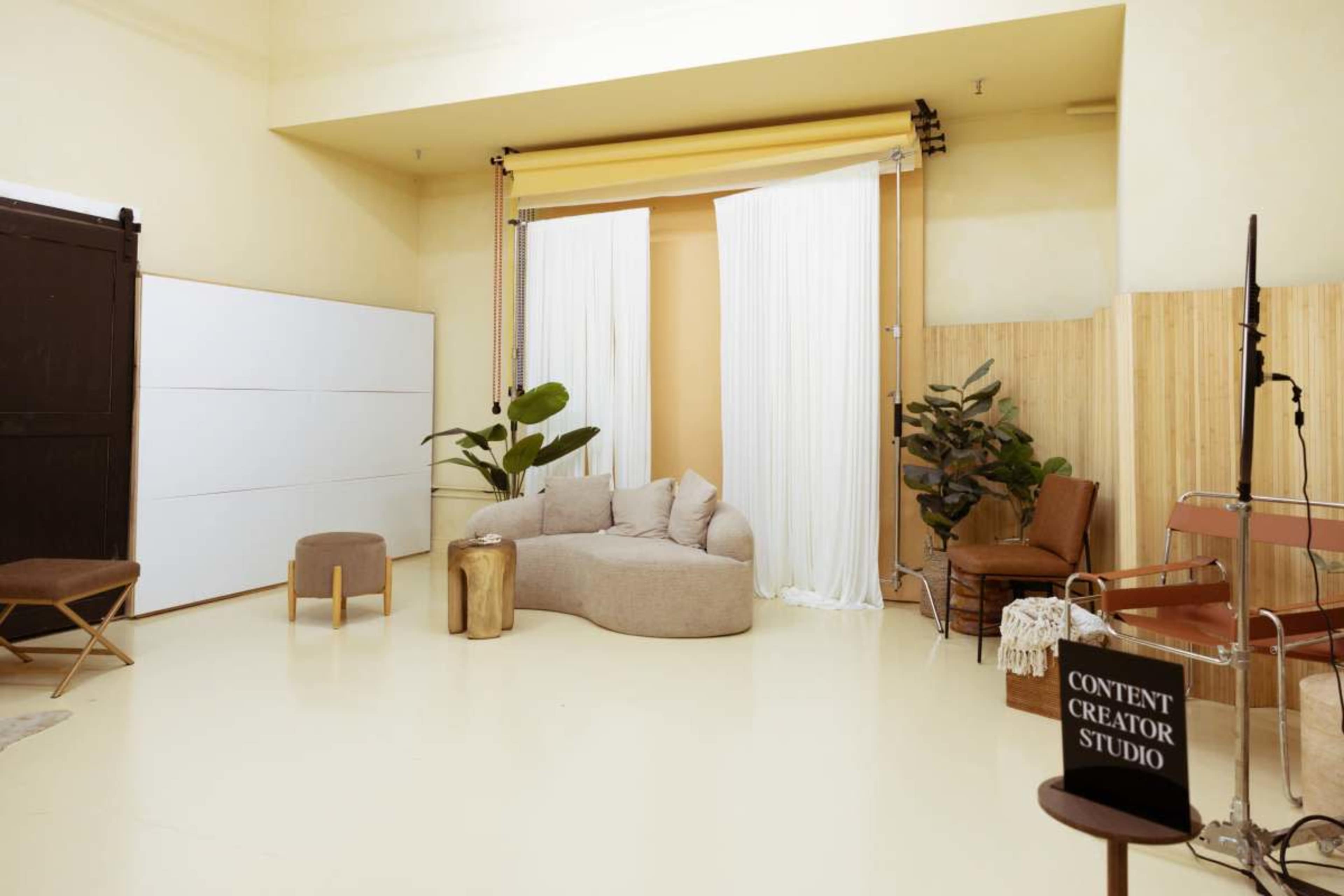 The image shows a minimalist content creator studio featuring a beige sofa, curtains, plants, and a sign that reads "CONTENT CREATOR STUDIO."
