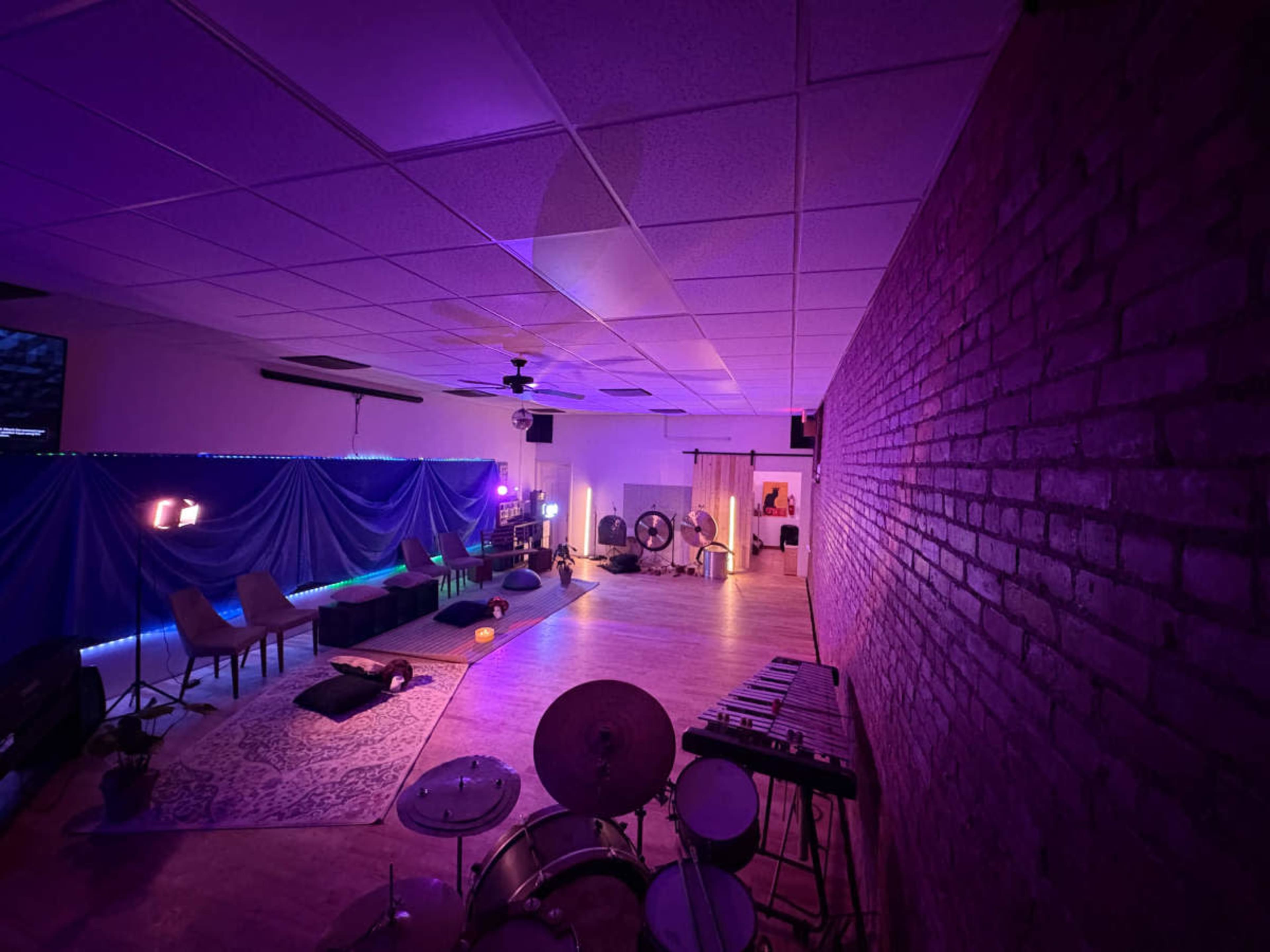 The image shows a spacious interior room with purple lighting, featuring seating areas, musical instruments, and decorative items arranged on the floor.
