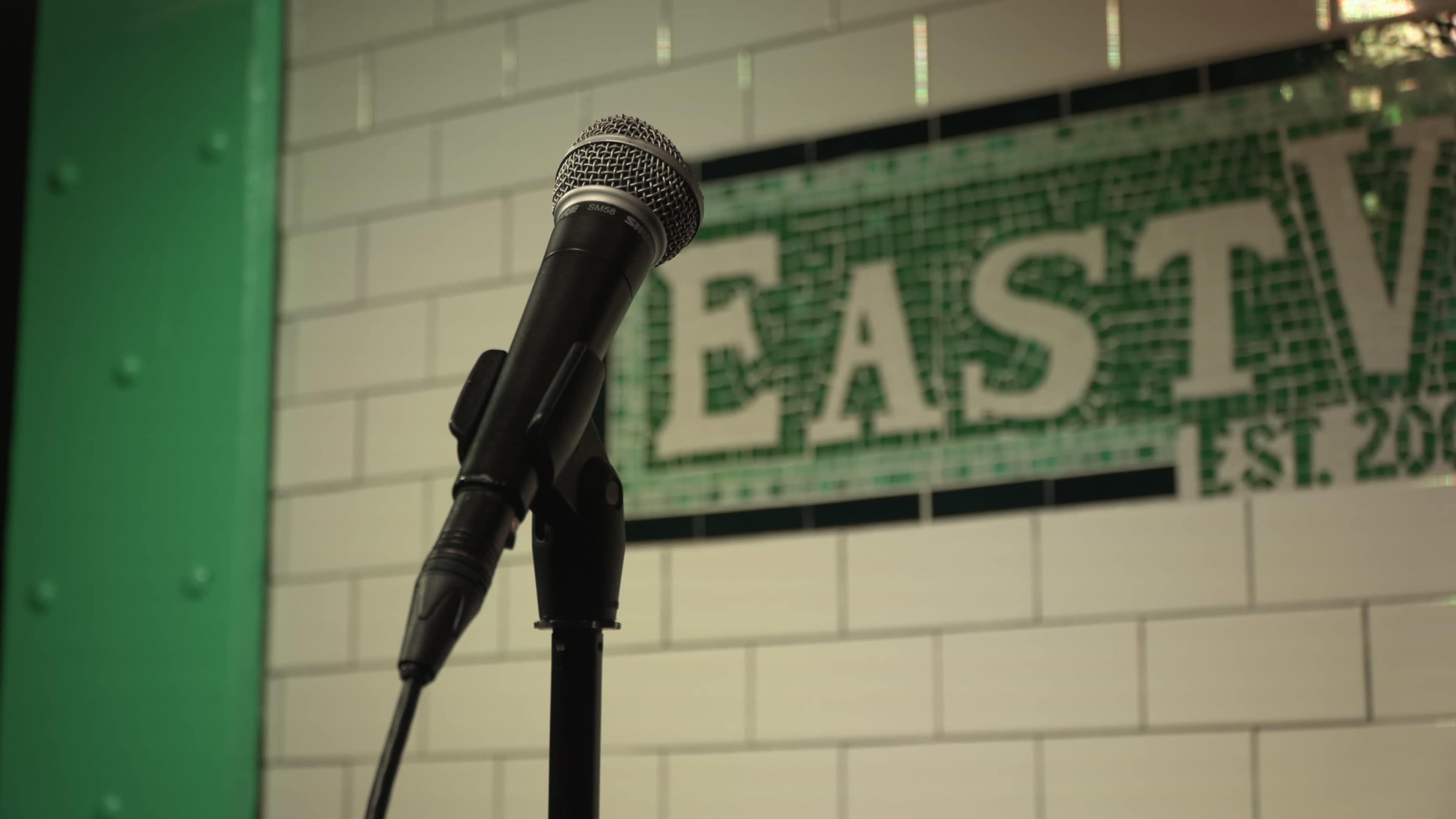 A microphone stands on a black stand in front of a tiled wall featuring the word "EAST" in green lettering.