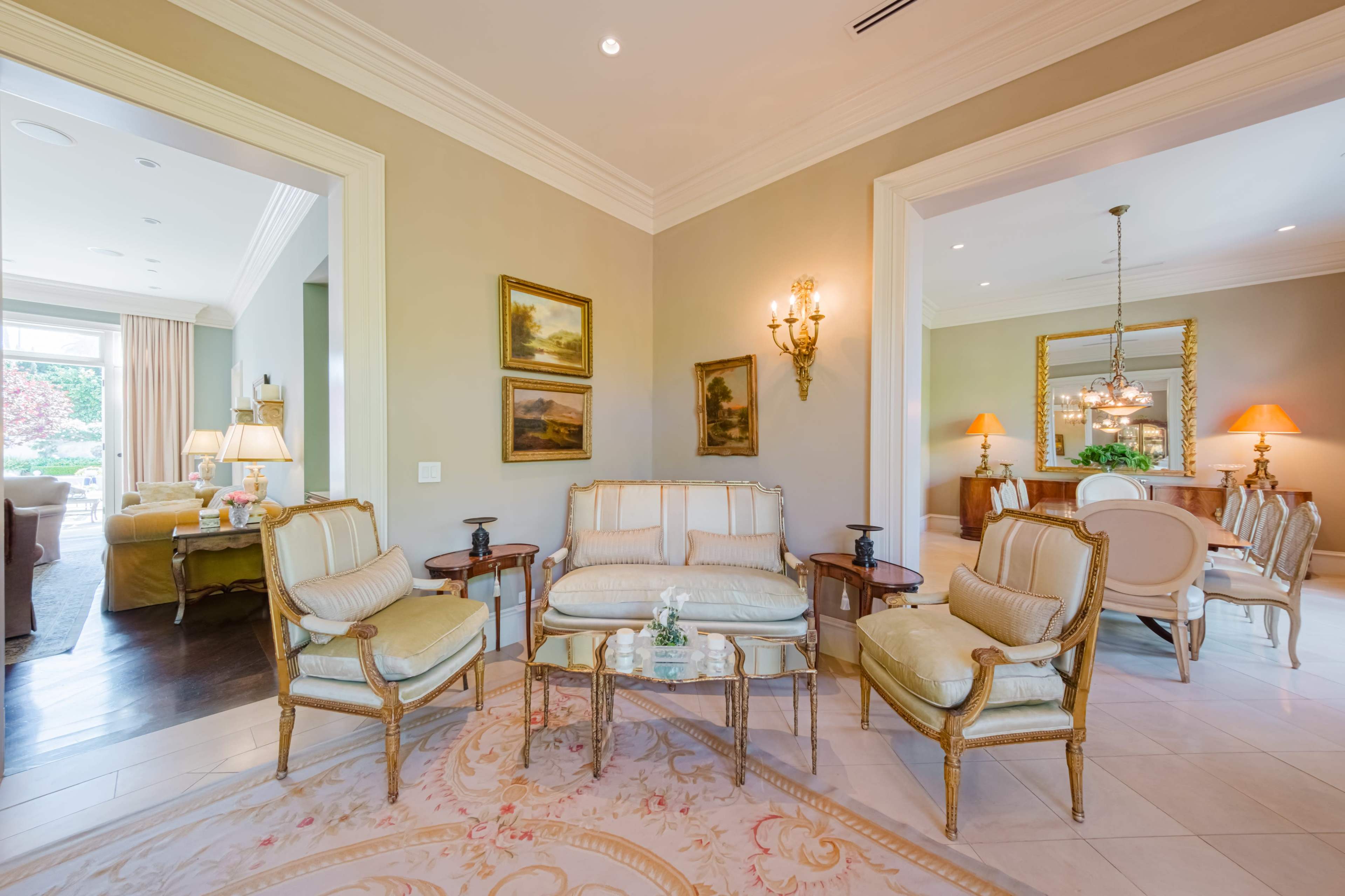 A formal living area with cream-colored walls, antique-style furniture, and framed paintings, connected to a dining area through open archways.