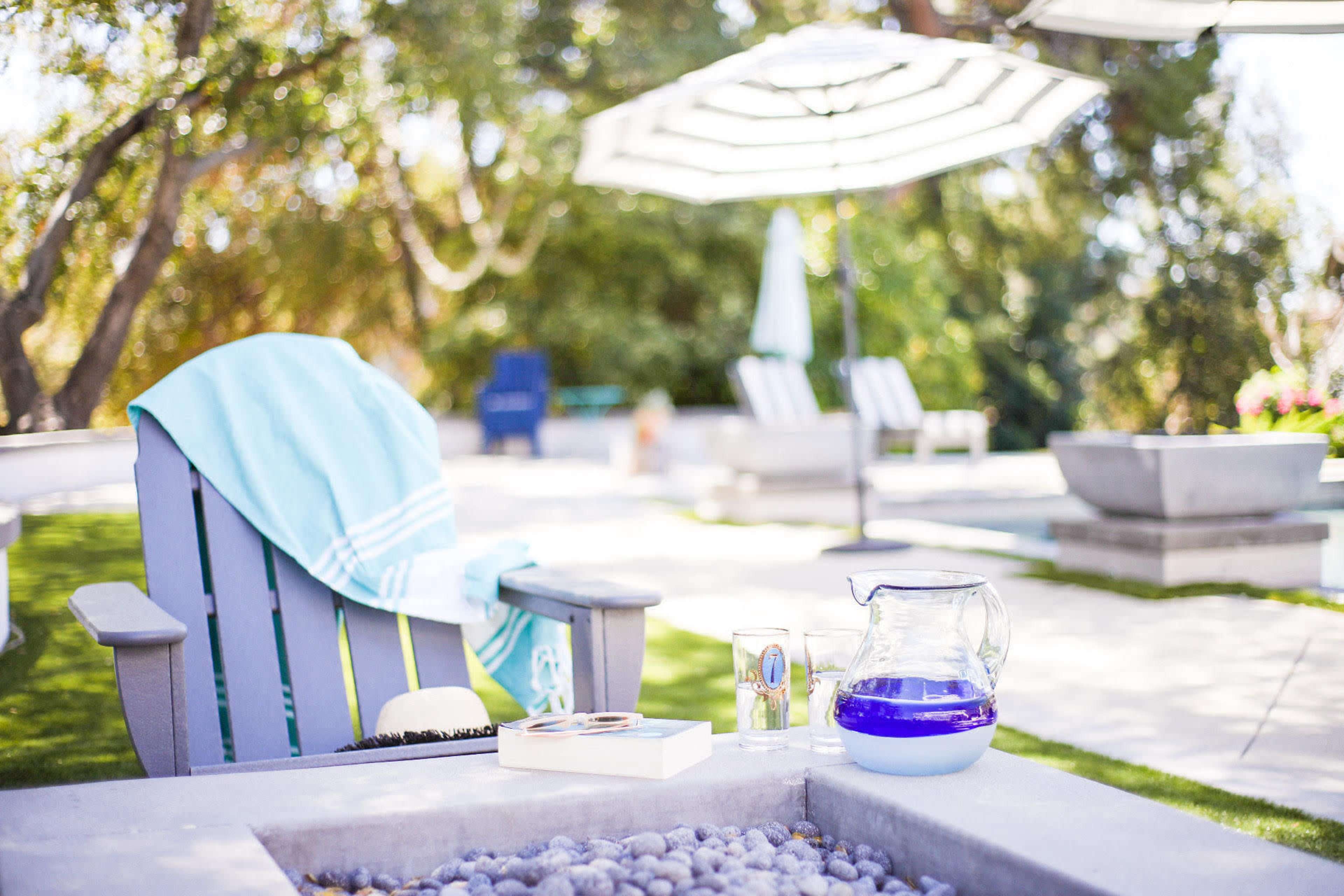 A patio scene features an Adirondack chair with a blue blanket, a glass pitcher, and a book near a fire pit, surrounded by greenery and outdoor seating under an umbrella.