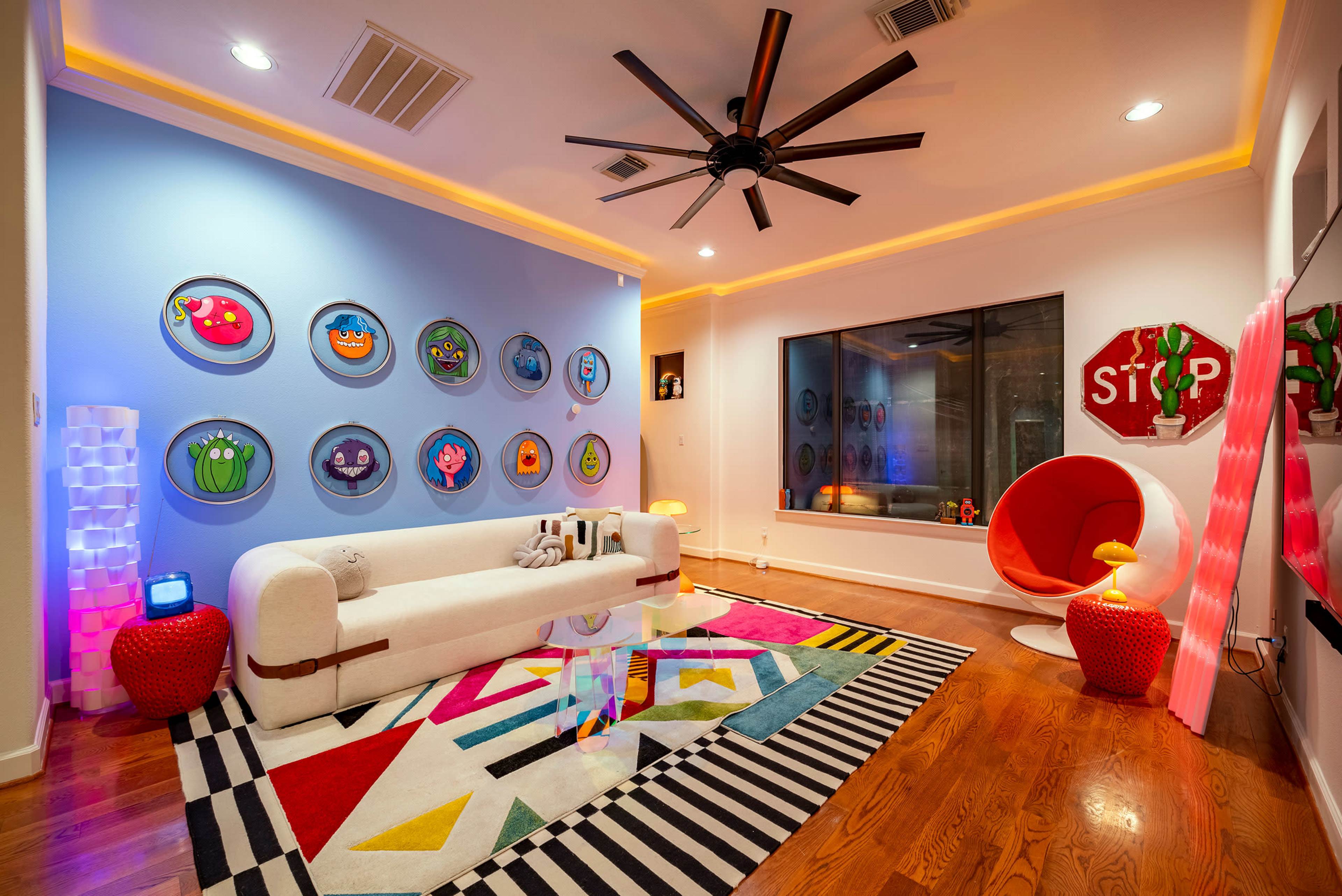 The room features a modern white sofa, colorful wall art, a circular red chair, and a patterned rug on hardwood flooring.
