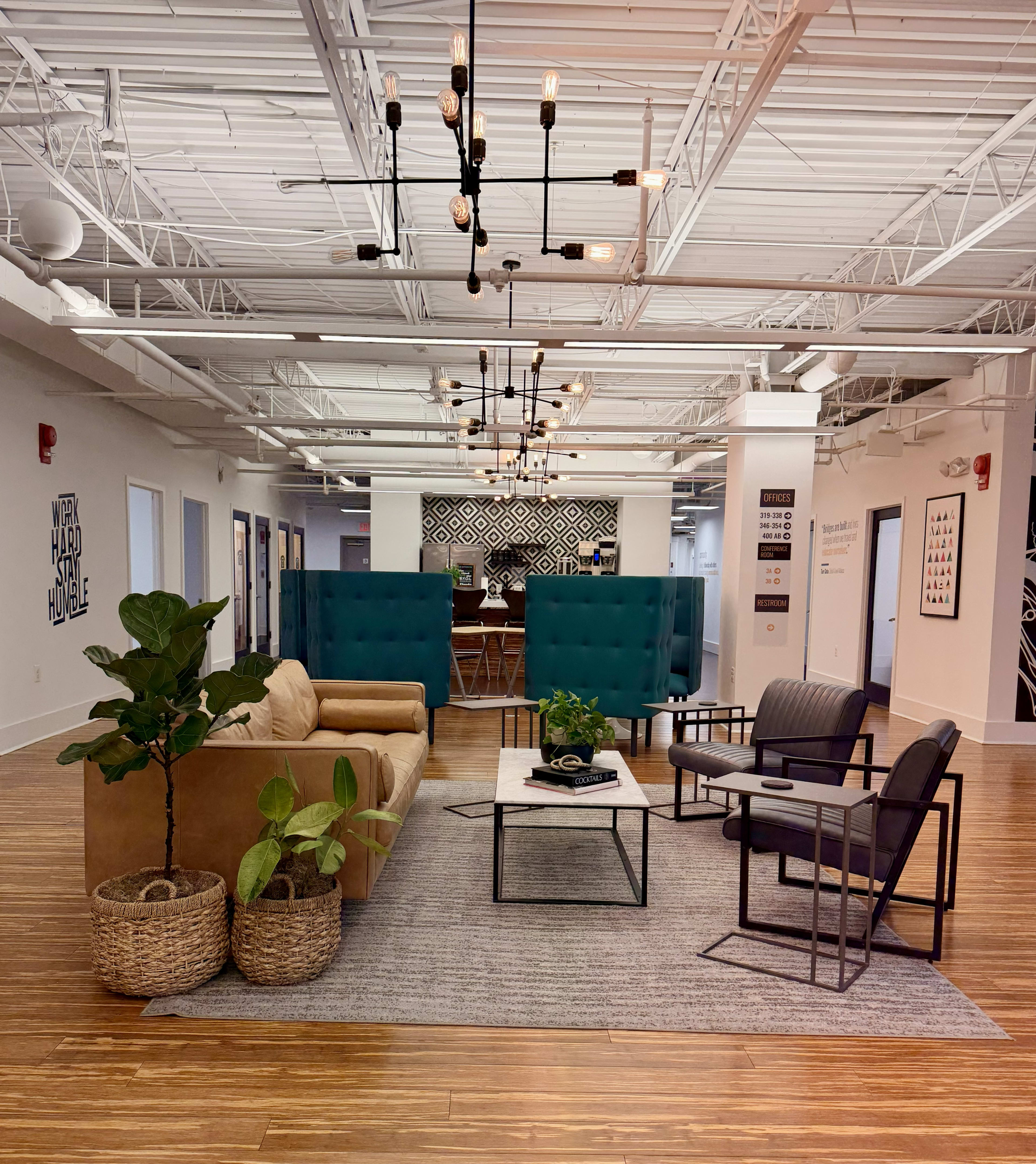 The image shows a modern office space with seating areas, decorative plants, and a large rug.