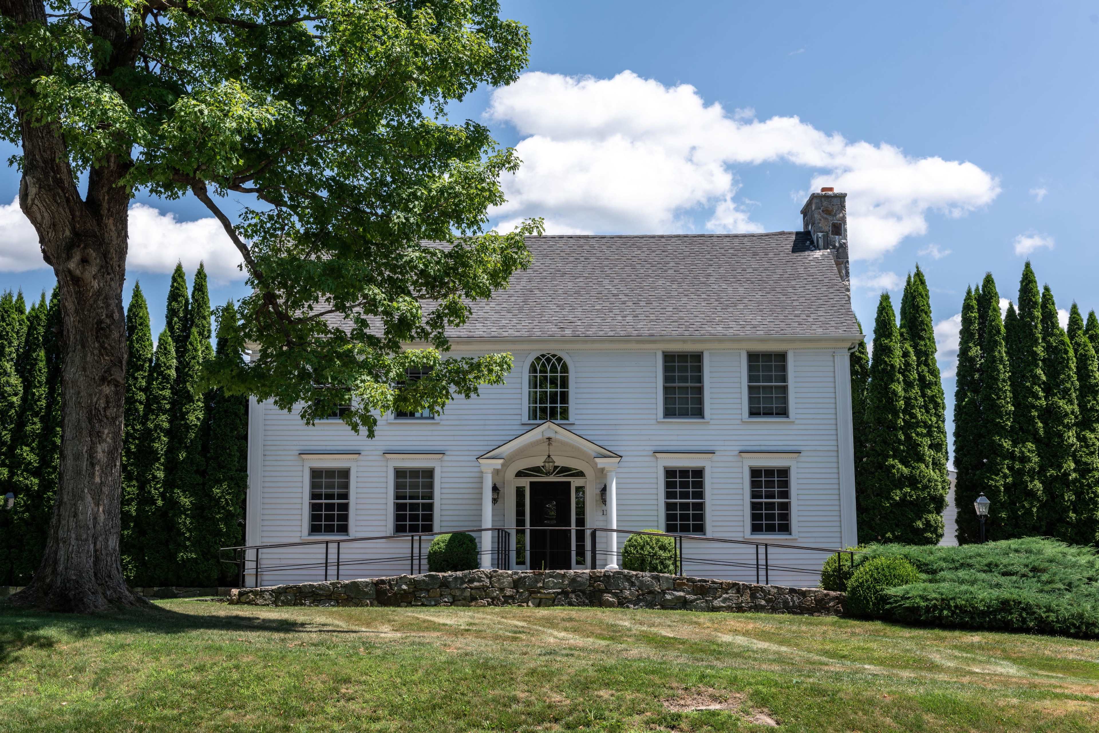 A two-story white house with a gray roof and a front porch is surrounded by well-maintained grass and tall evergreen trees.