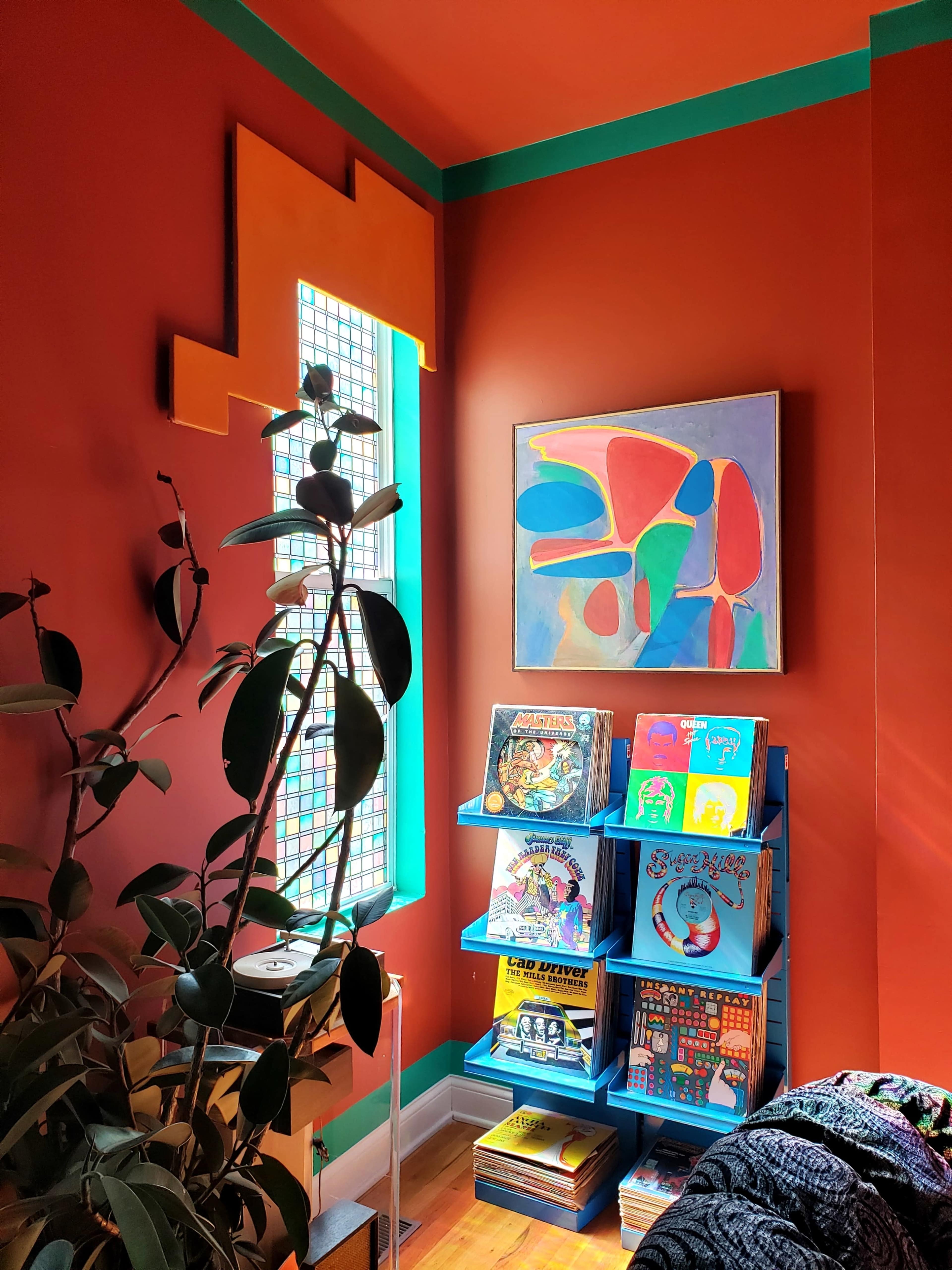 A brightly colored corner of a room features a tall plant, a blue bookshelf filled with vibrant books, and an abstract painting on the wall beside a decorative window.