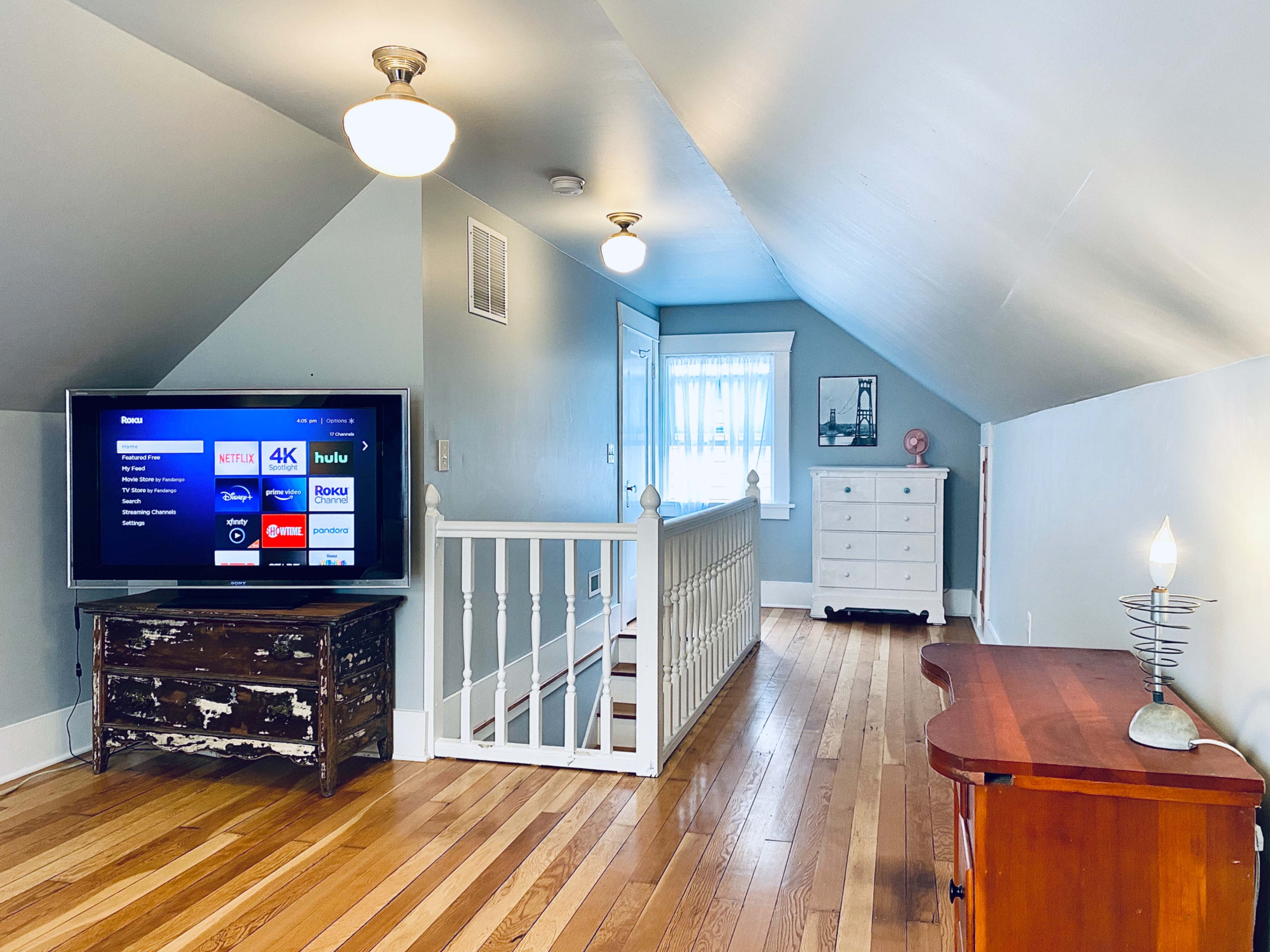 The image shows an attic space with a television on a distressed wooden table, a white dresser, and a small table with a lamp, all under a sloped ceiling.