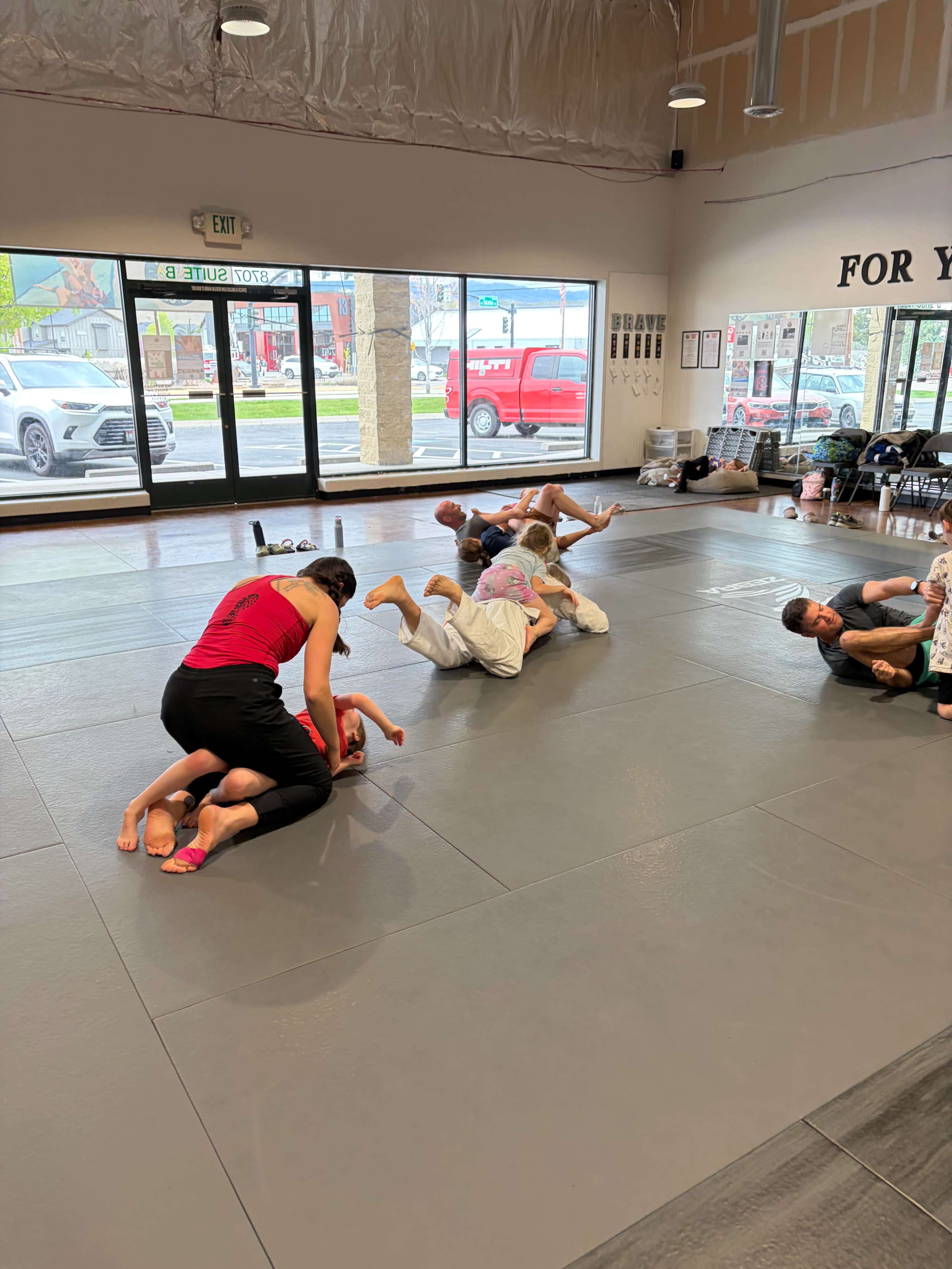 A group of participants is engaged in a grappling training session on a gray mat in a brightly lit martial arts studio.
