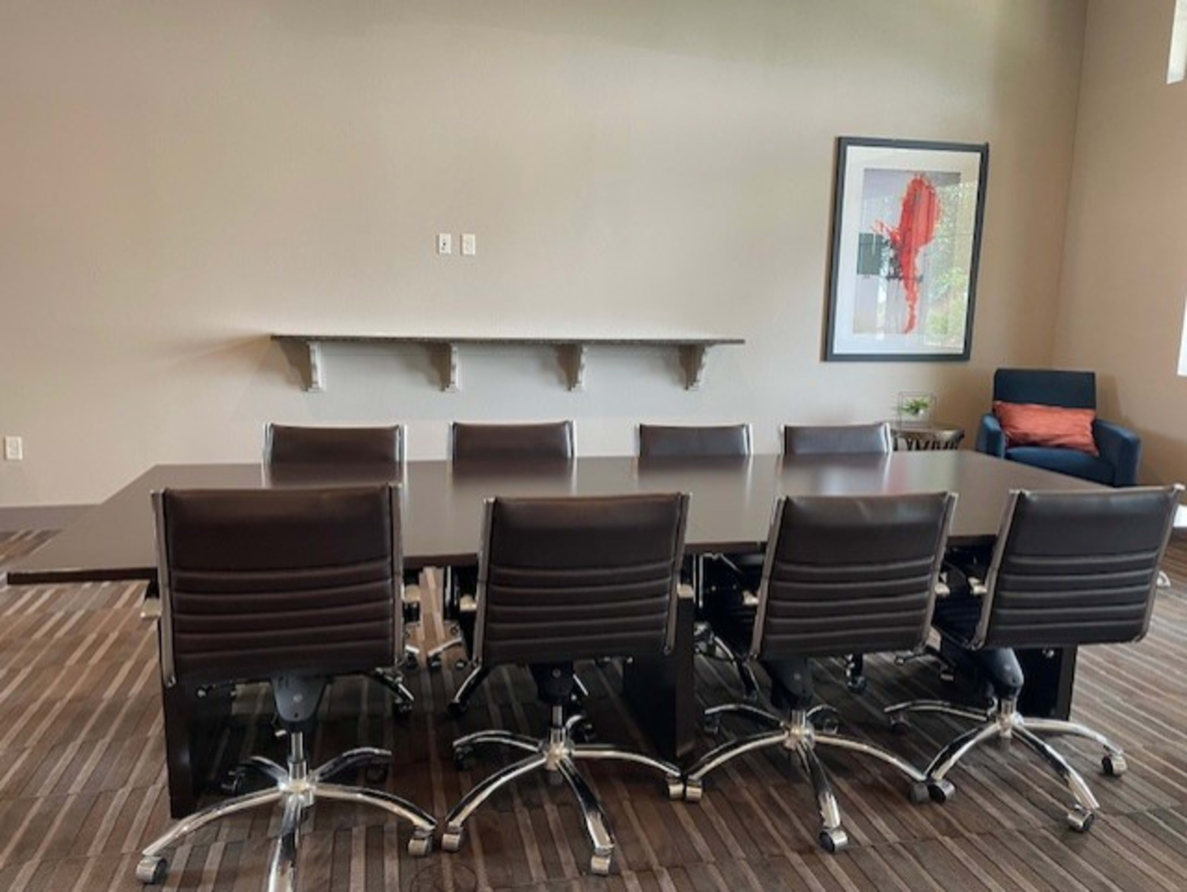 A large conference table with eight chairs is positioned in a meeting room featuring a minimalist design and a wall-mounted shelf.