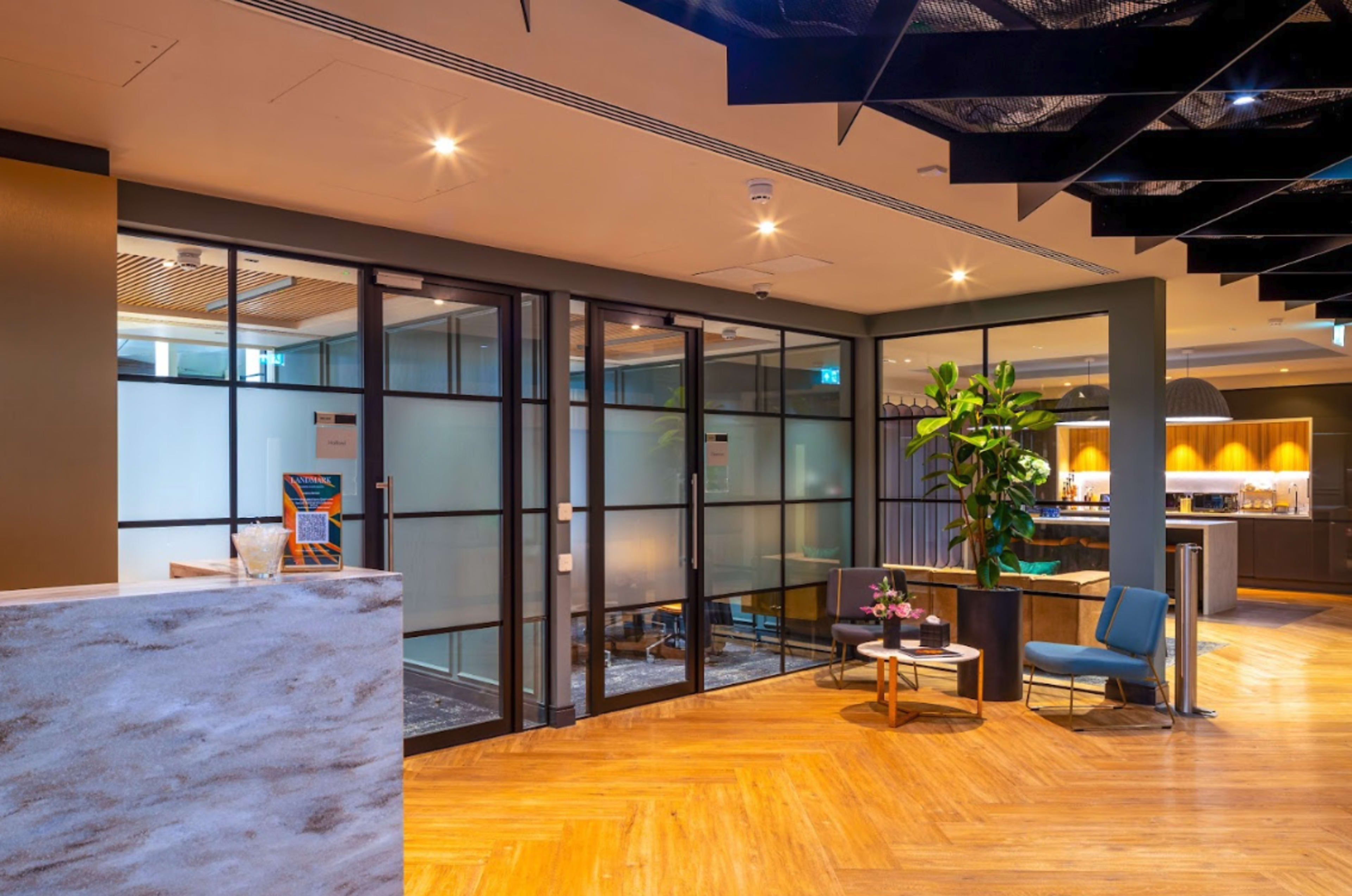 A modern office lobby with glass partitioned meeting rooms, a reception desk, and a seating area featuring a plant and a small table.