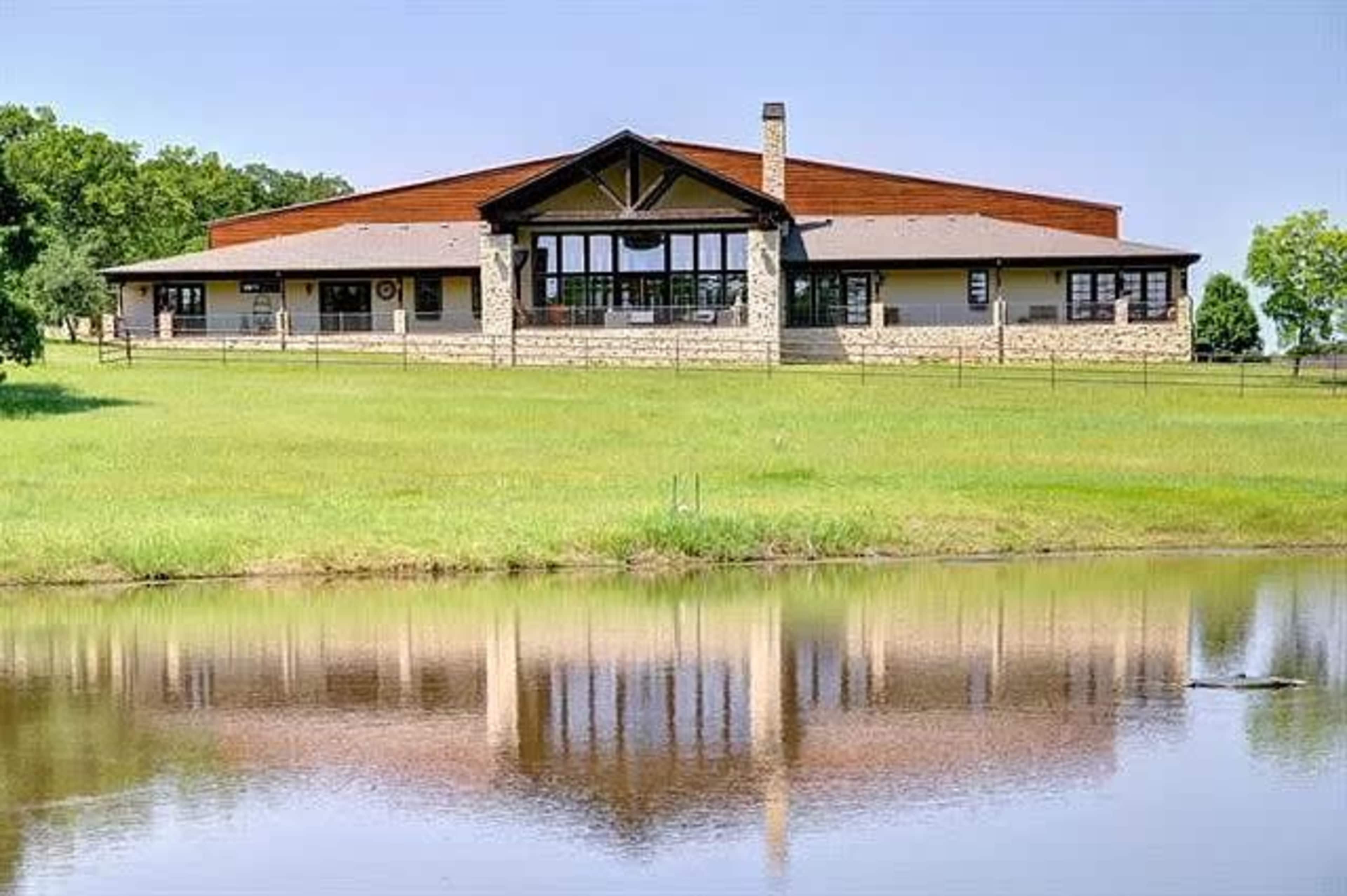 A large ranch-style building with a stone base and a pitched roof, reflected in a calm pond surrounded by green grass.