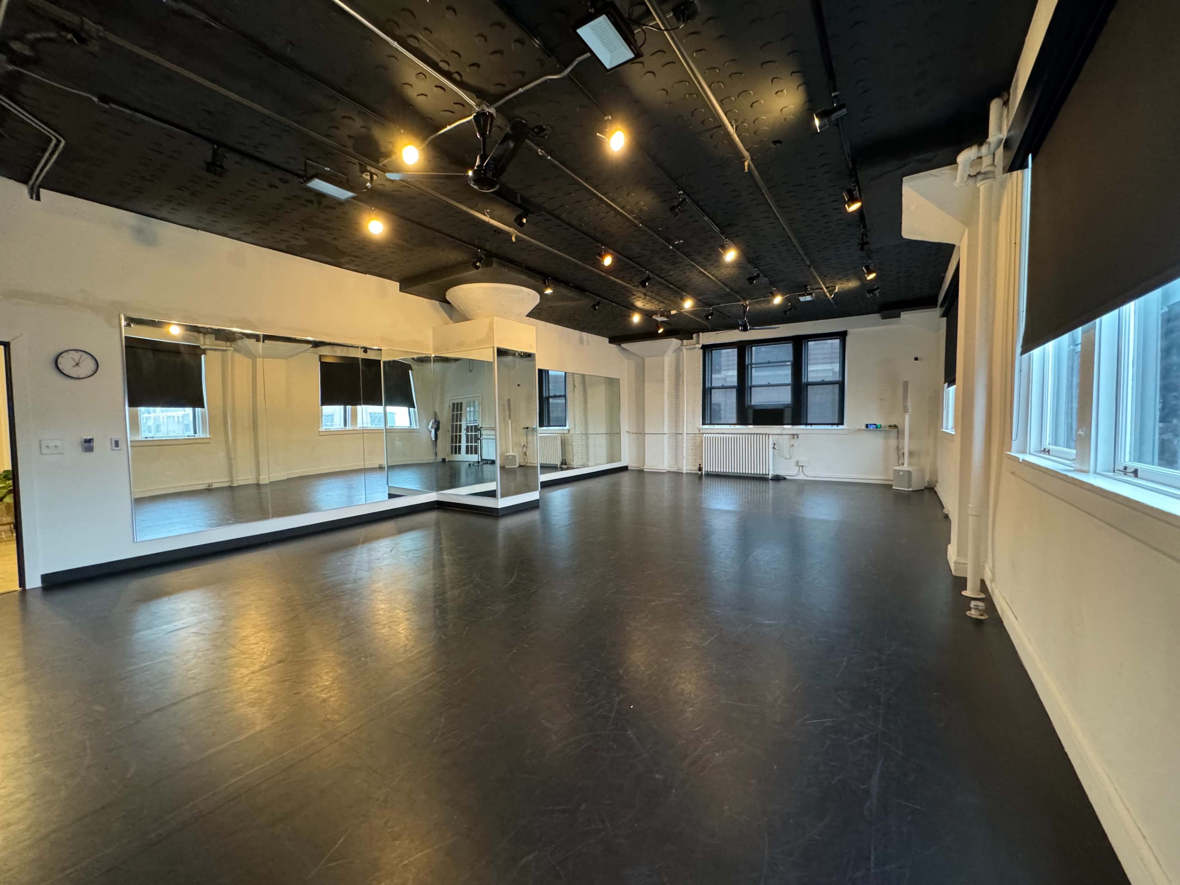 A spacious dance studio with black flooring, large mirrors on one wall, and ample natural light from windows.