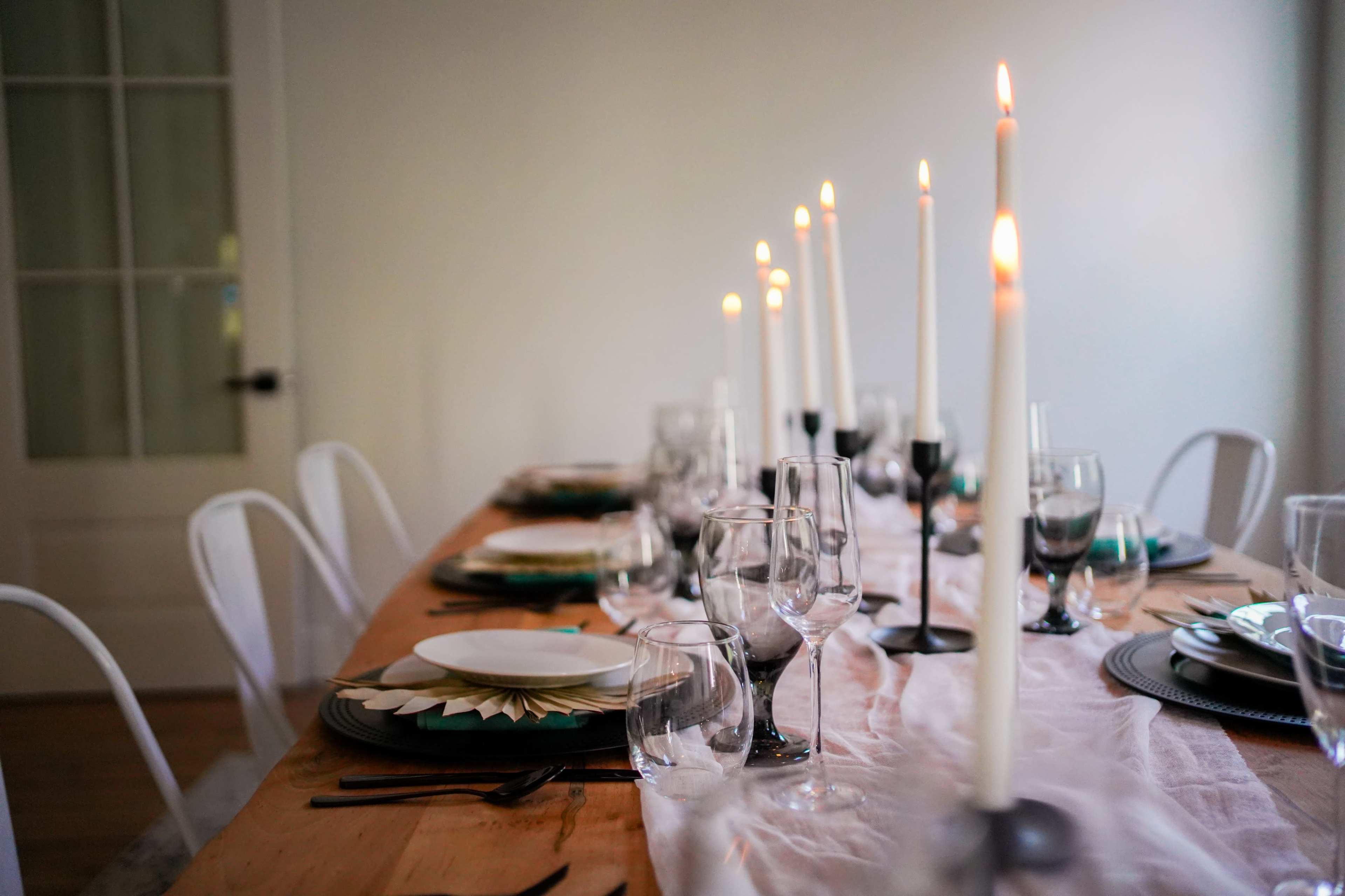 A long wooden dining table is set with plates, glasses, and lit candles arranged in a row.