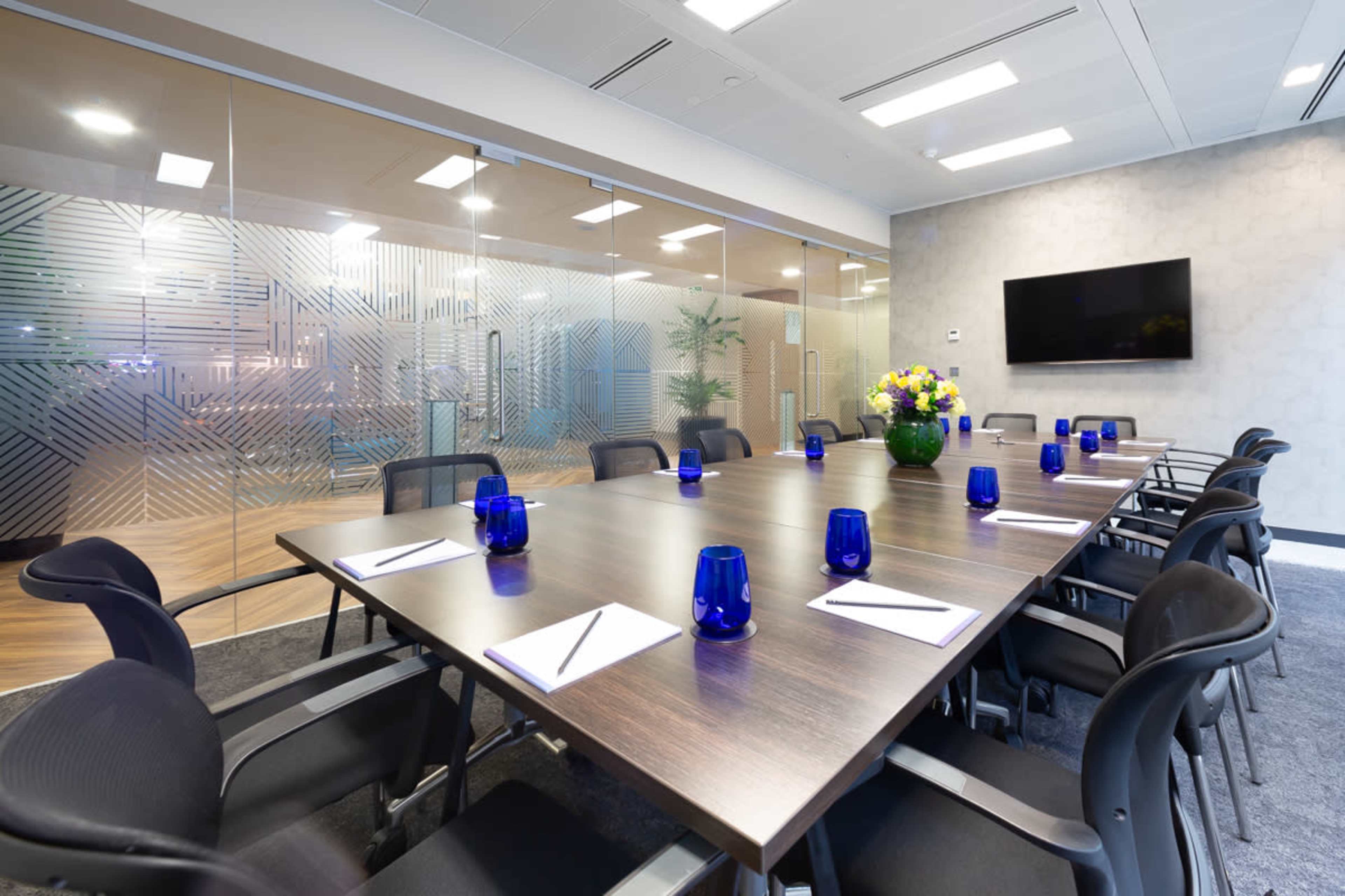 A modern conference room features a large wooden table surrounded by black chairs, with blue glass candles and notepads set at each place, and a wall-mounted screen visible in the background.