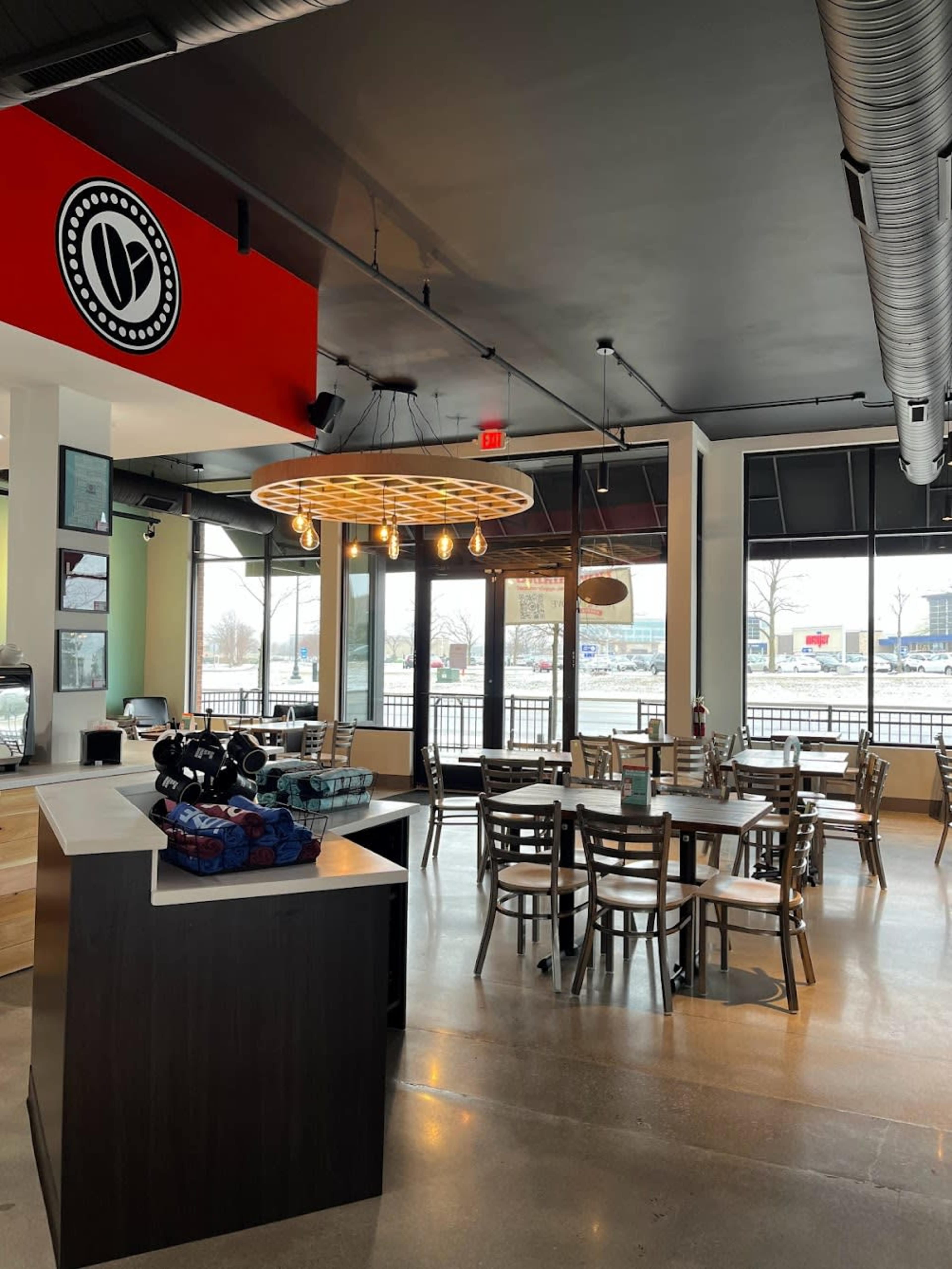 A modern café interior with a round light fixture, wooden tables and chairs, and large windows overlooking a view outside.